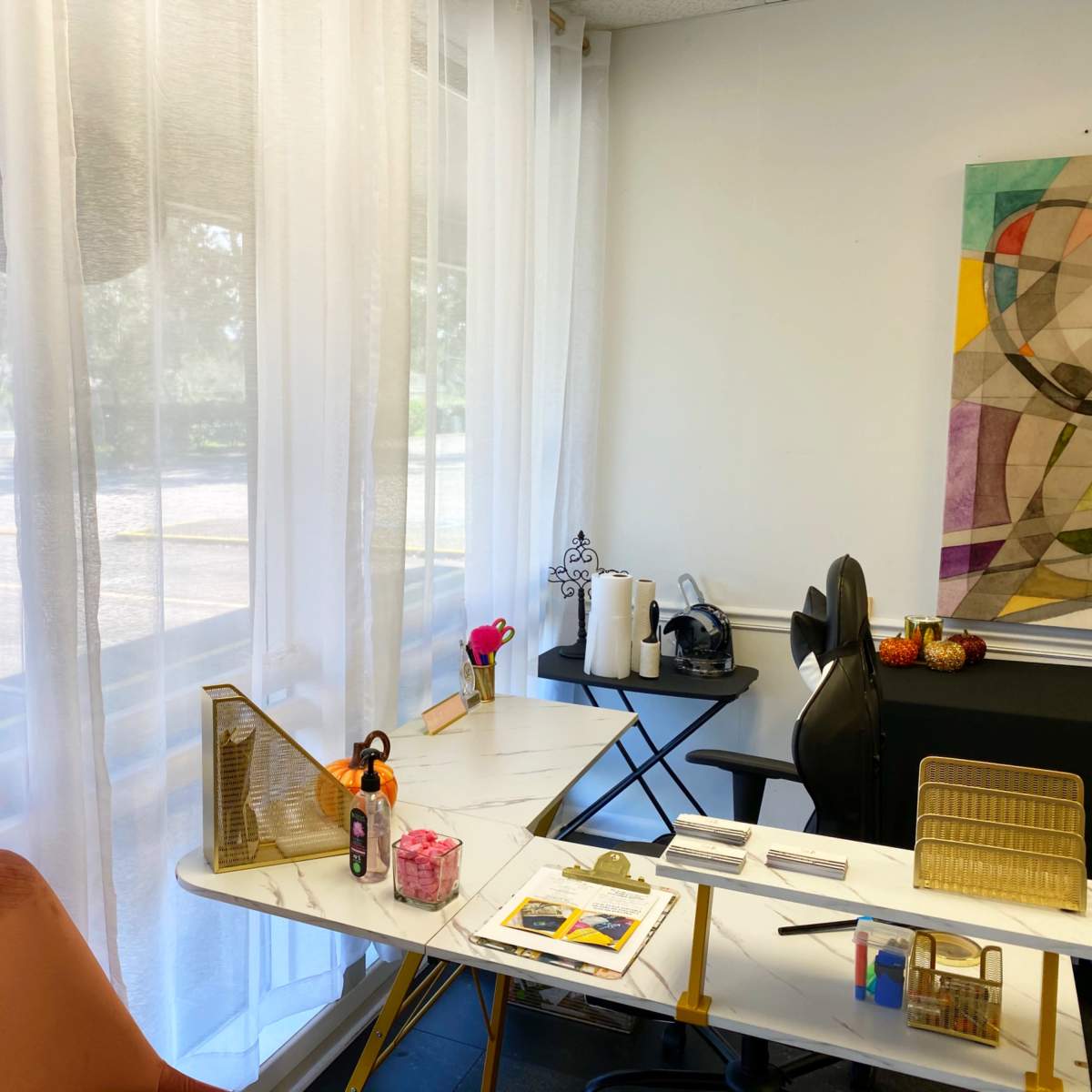 A bright room features a desk with art supplies, a chair, and sheer curtains allowing natural light to filter through.