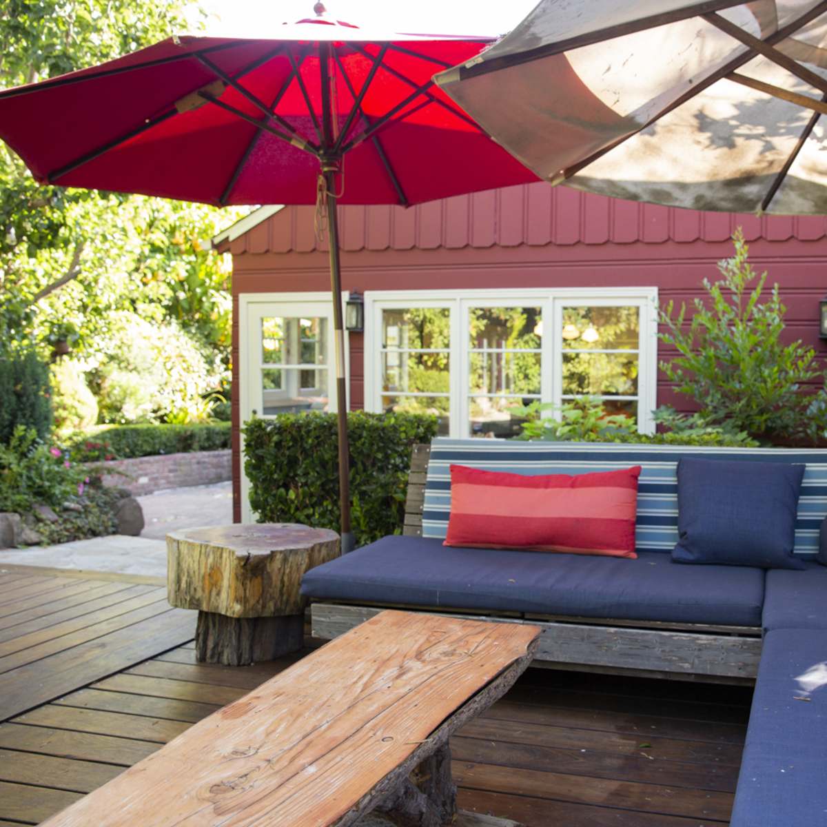 A backyard seating area features striped sofas, red umbrellas, and a wooden table.