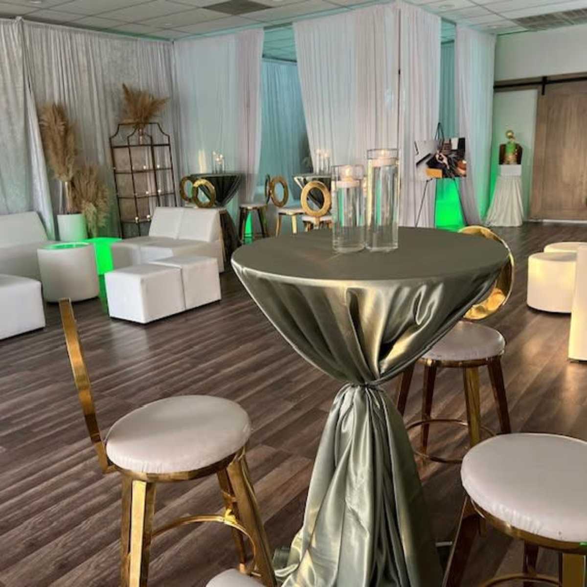 A decorated event space with white drapes, modern seating, and a table draped in a greenish cloth with candles.
