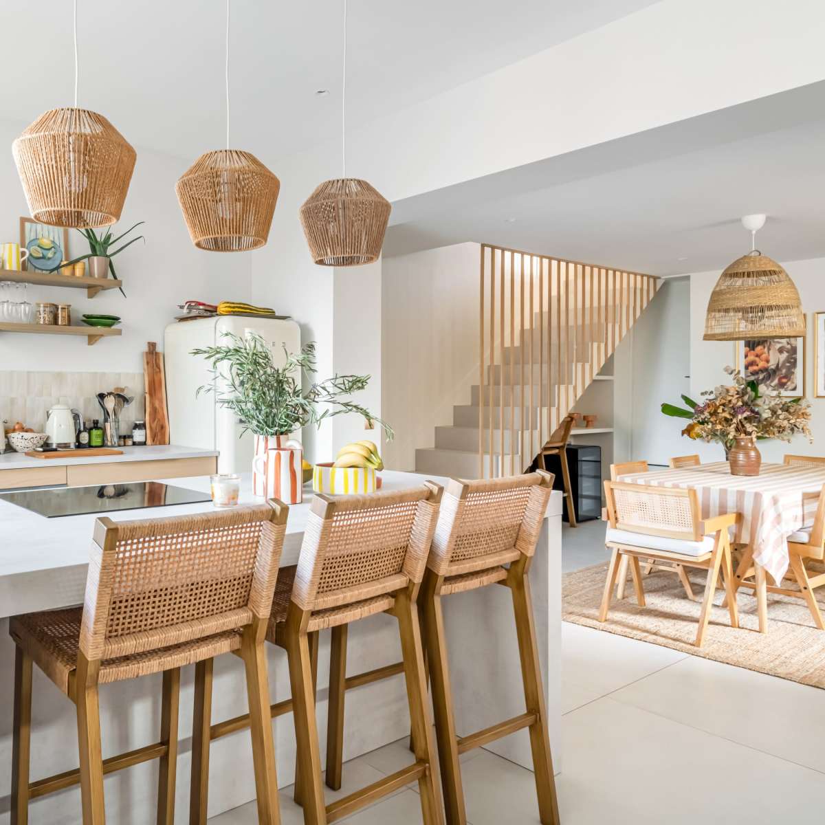 A modern kitchen features light wood cabinetry, woven pendant lights, and a dining space with a table set for six.