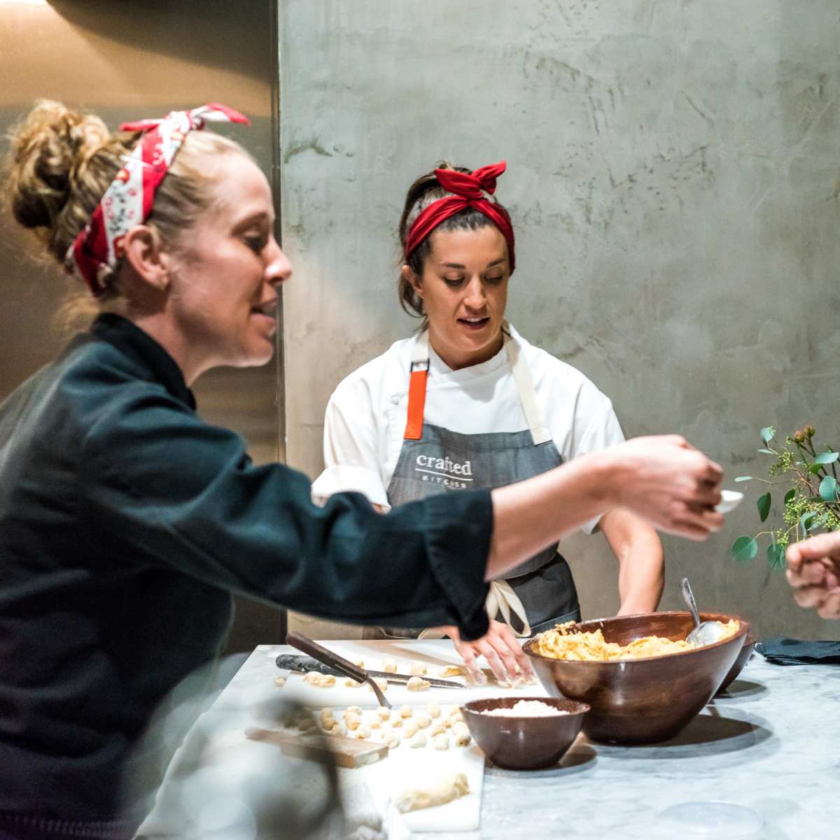 Three people are interacting in a kitchen, with two women preparing food and a man observing while holding a small dish.