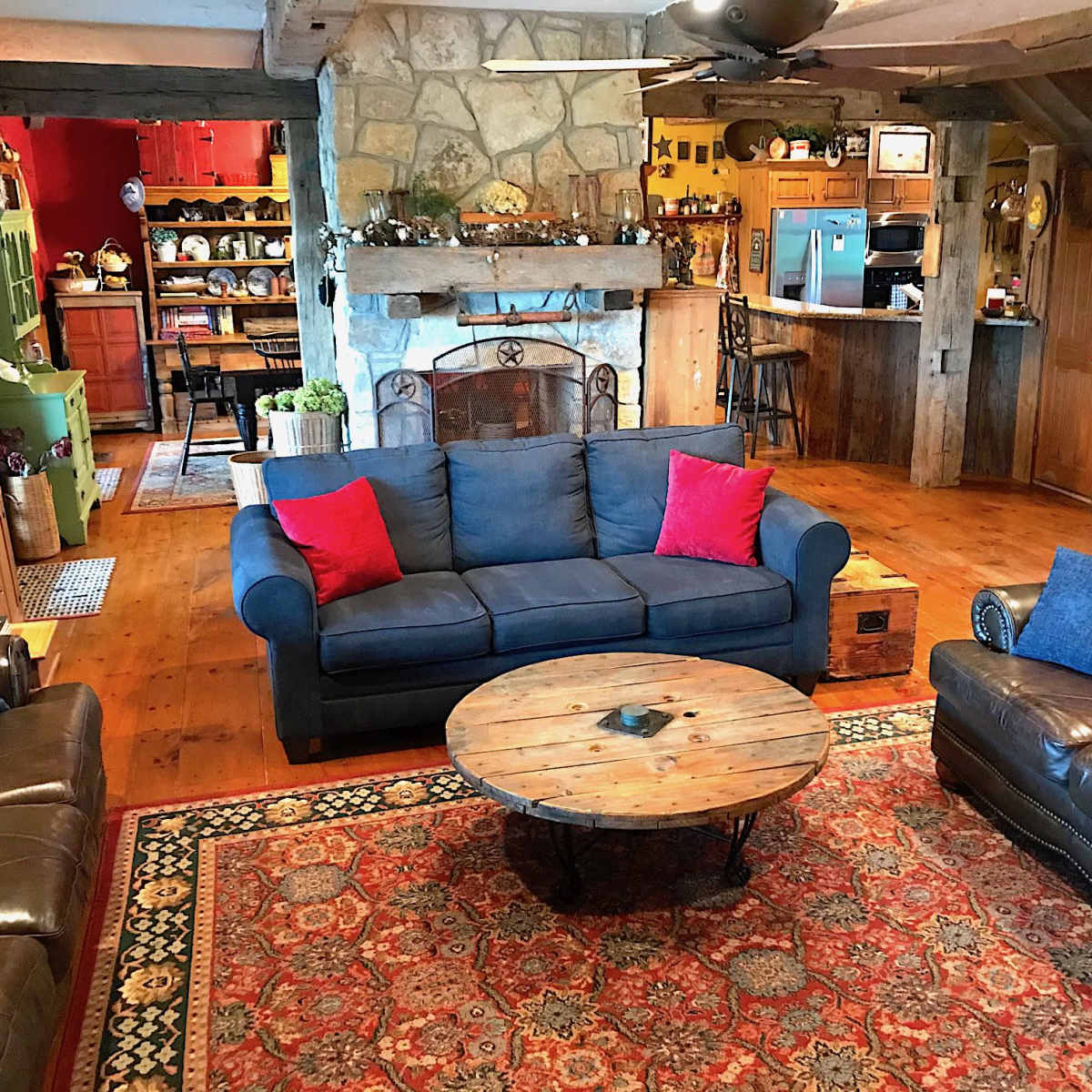 A cozy living room features a blue sofa with red cushions, a round wooden coffee table, and a stone fireplace, surrounded by wooden beams and rustic decor.