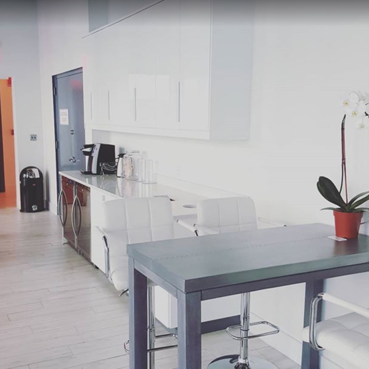 A modern kitchen area with white cabinetry, a coffee station, and a small dining table with bar stools.