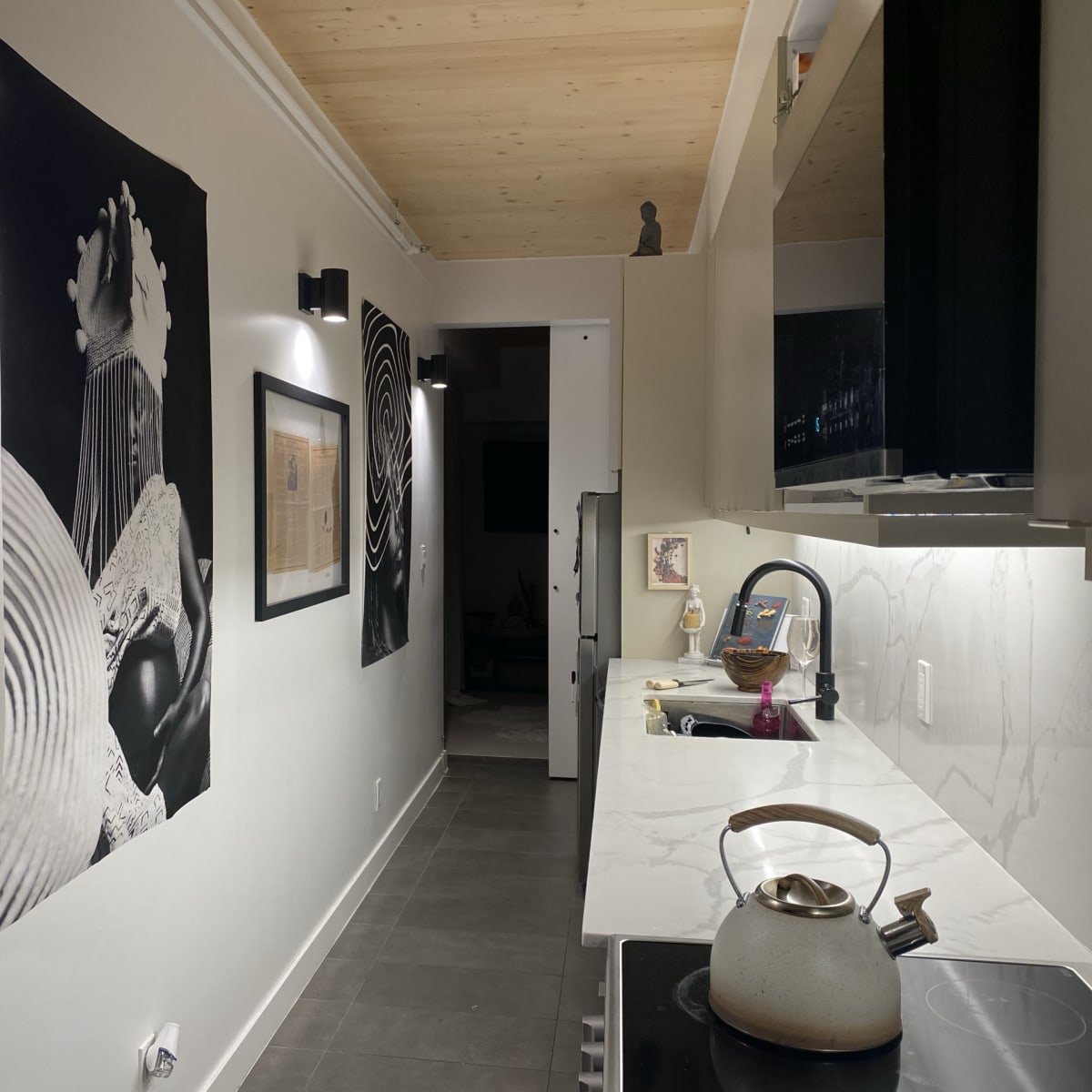 A narrow hallway features black-and-white artwork on the walls, with modern cabinetry and a kettle on a stove in the kitchen area.