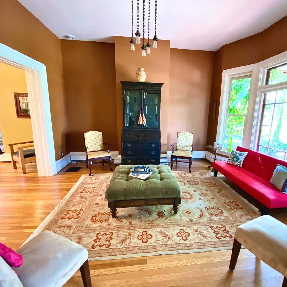 A living room with a red sofa, four chairs arranged around a green ottoman, and large windows allowing natural light to enter.