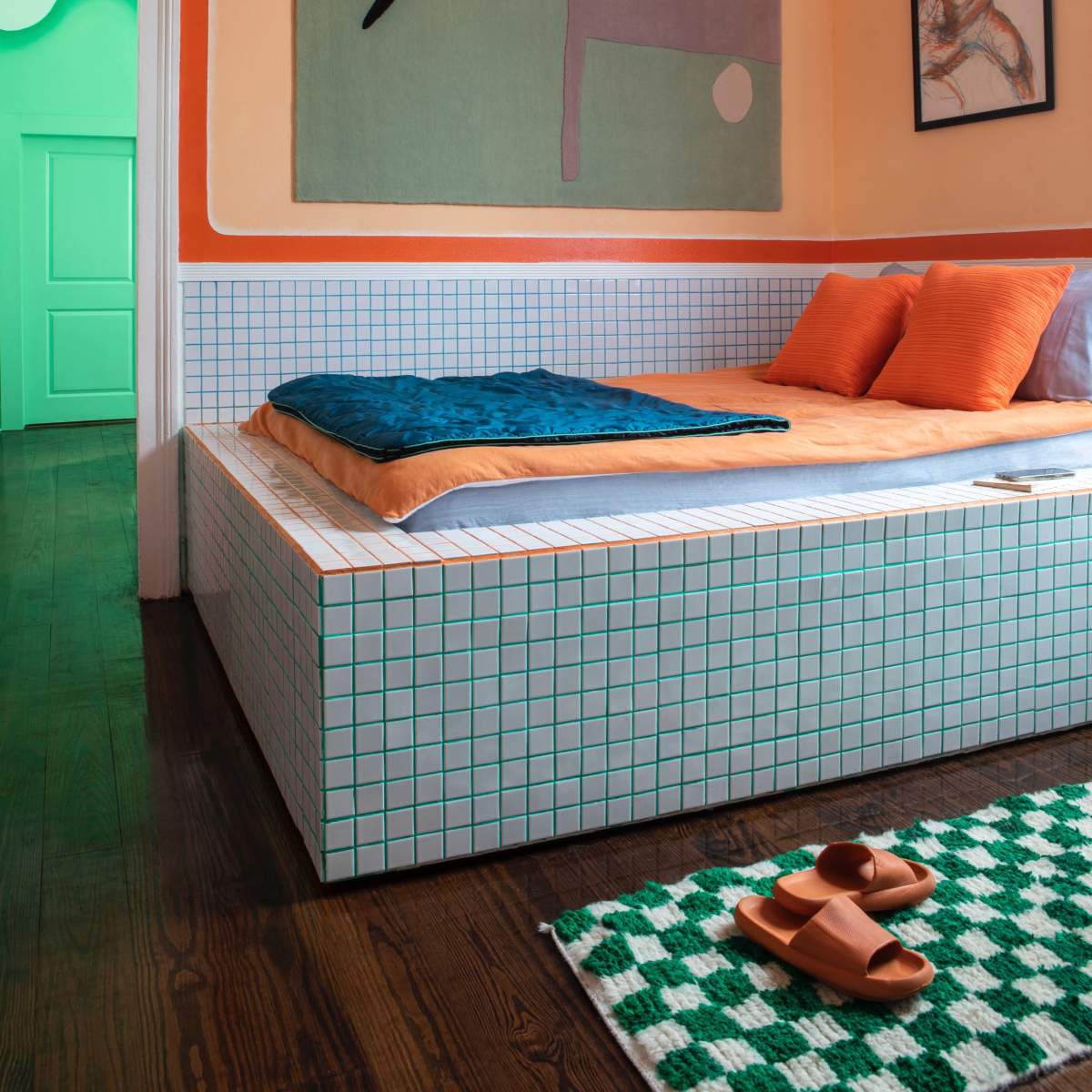 A modern bedroom with a tiled bed frame, orange walls, and colorful decor, including a large artwork and a round mirror pendant.