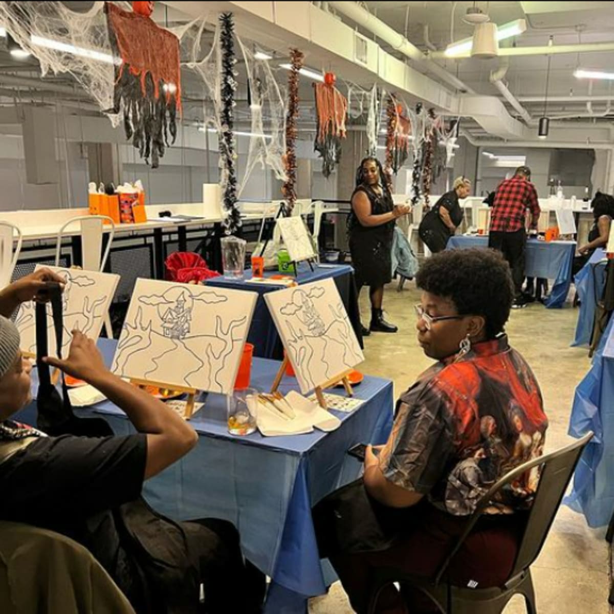 Two individuals sit at a table adorned with canvases and art supplies in a decorated room featuring Halloween-themed decorations and people engaged in painting activities.