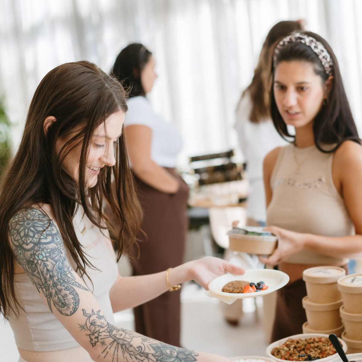 A woman with tattoos selects food from a buffet while another woman stands nearby, holding a phone.