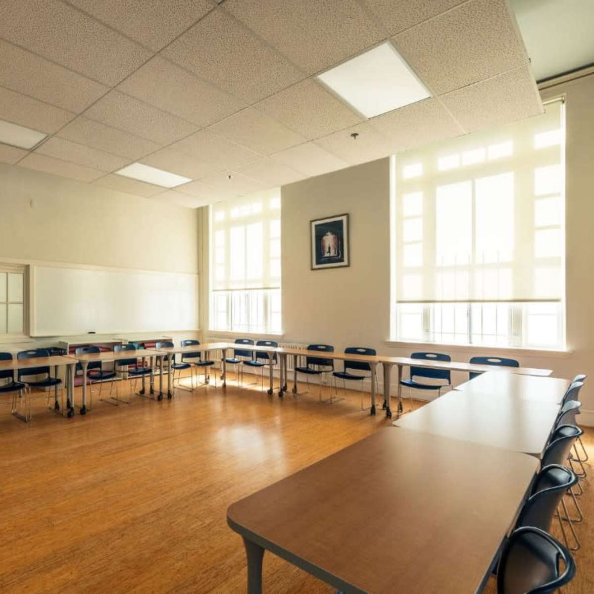 A bright classroom with wooden flooring, several tables arranged in a U-shape, and large windows allowing natural light to enter.