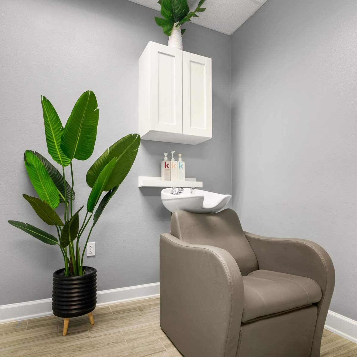 A small salon corner featuring a gray chair, a white cabinet mounted on the wall, and a potted plant in a black pot.