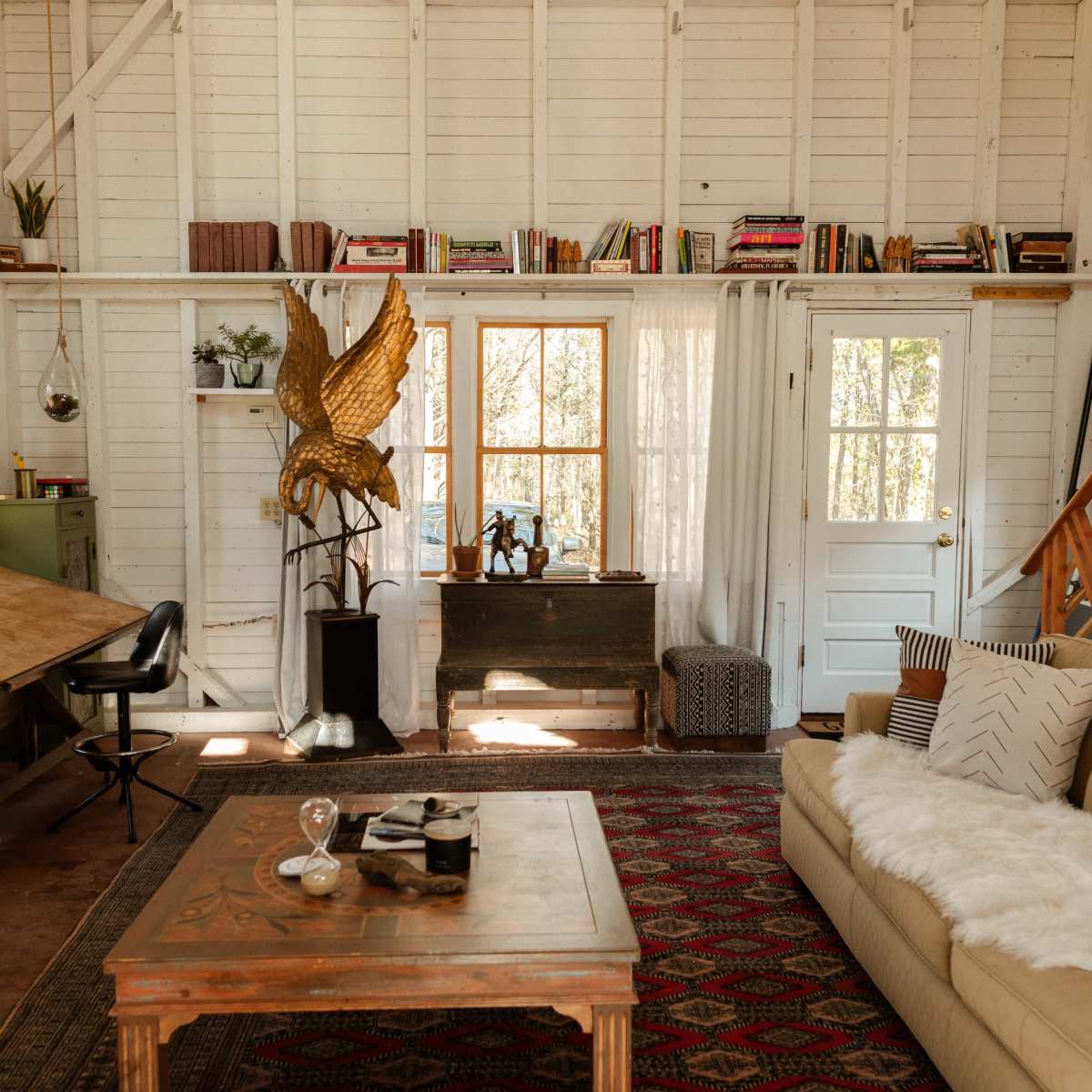 A cozy living space with a beige sofa, a wooden table for work, and bookshelves lined with books, accented by a golden sculpture and a large window allowing natural light.