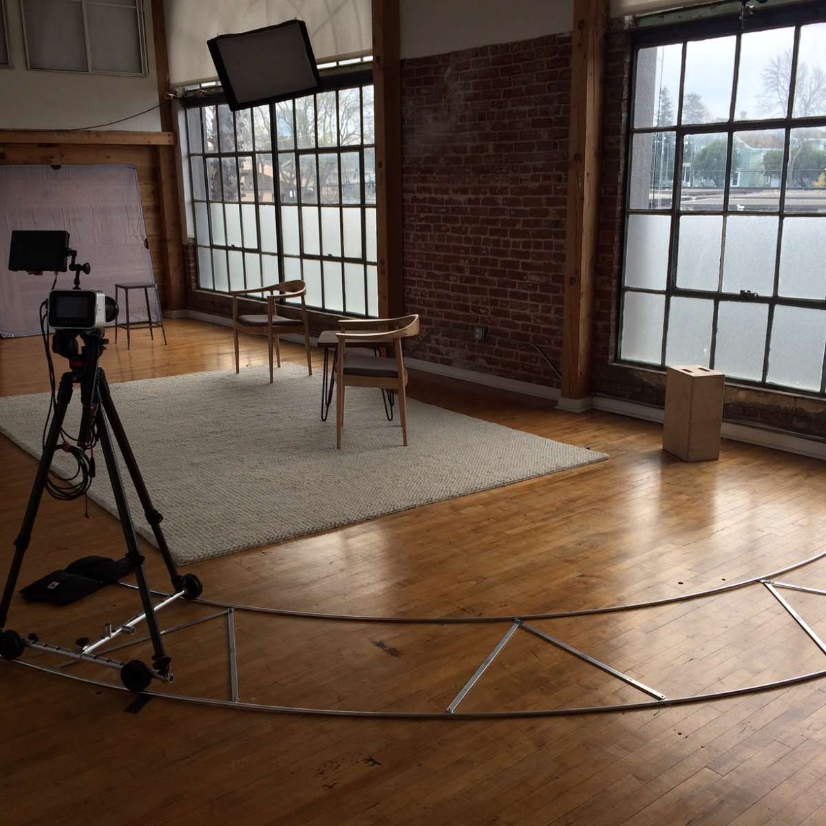 A film set featuring a circular camera track, a tripod with a camera, two chairs, and large windows letting in natural light.