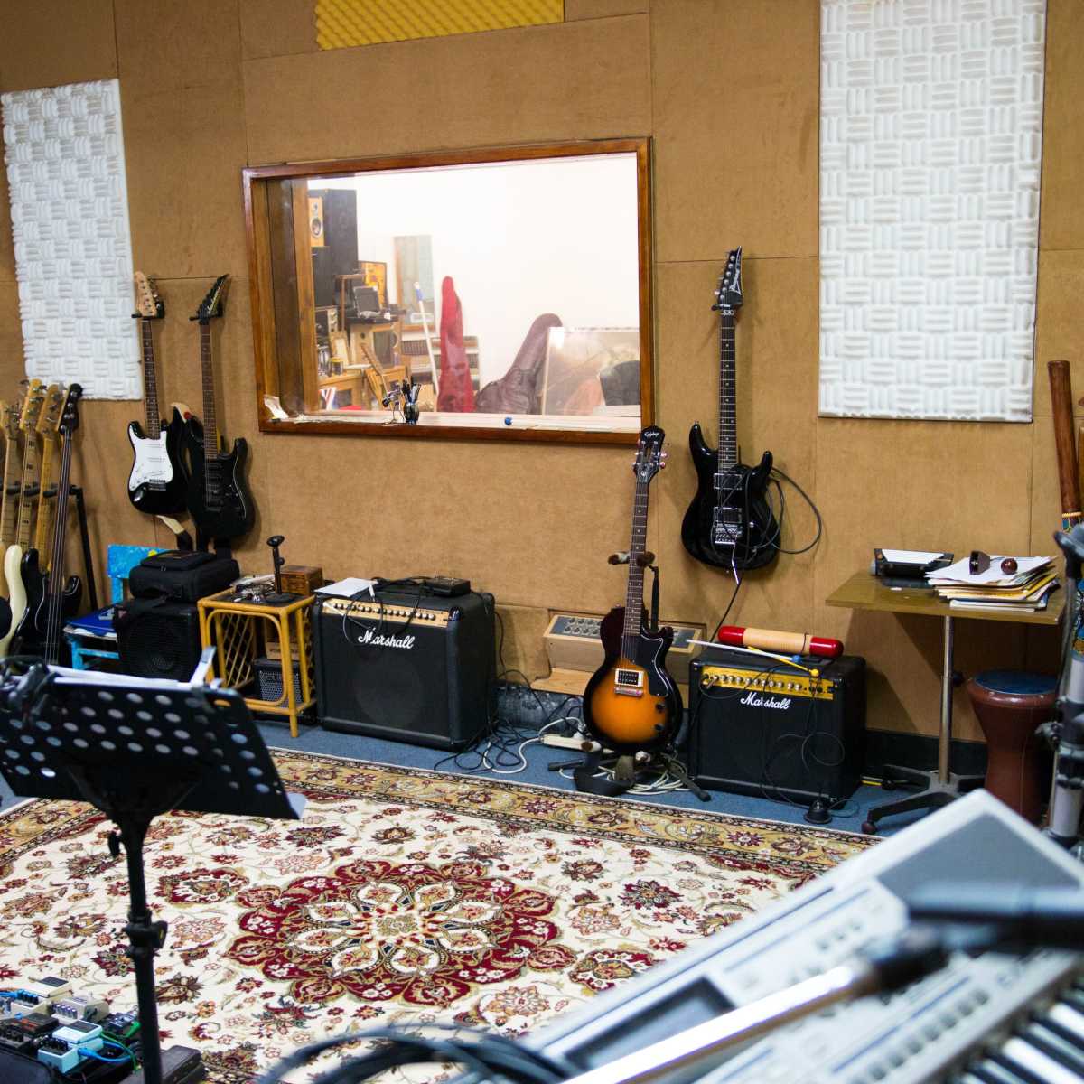 A music recording studio with various guitars, amplifiers, and a patterned rug, along with a control room visible through a window.