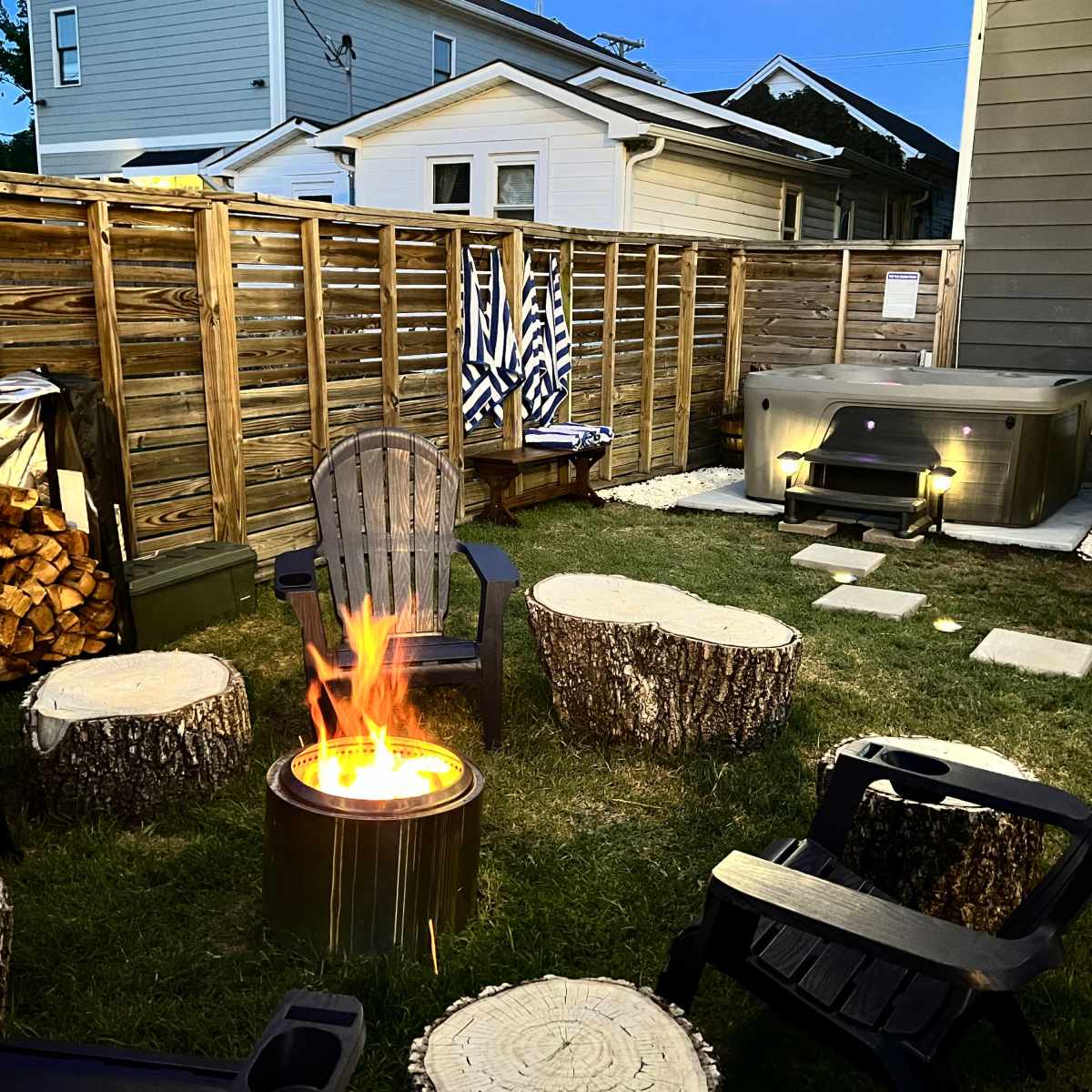 A backyard with a fire pit surrounded by wooden stump chairs, a hot tub in the corner, and a fenced area with stacked wood and a few decorative items.