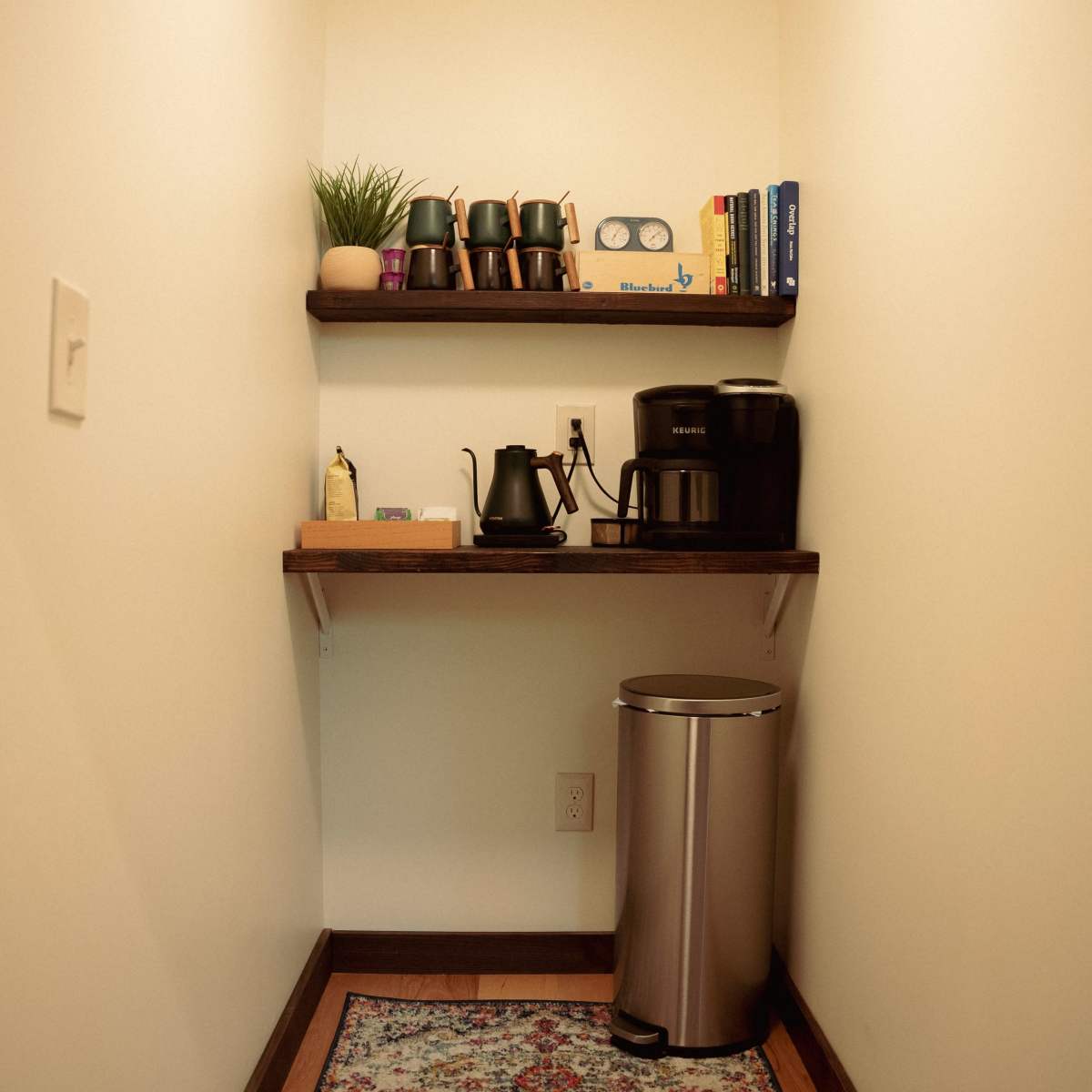 A small nook features a coffee station with shelves holding cups and books, alongside a coffee maker and trash can.