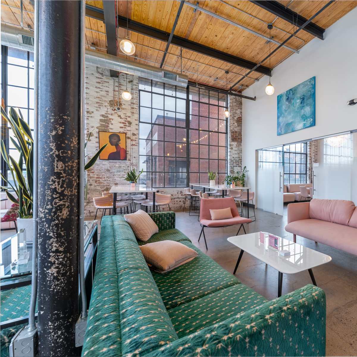 A modern, industrial-style lounge area featuring a green couch, pink sofa, and large windows allowing natural light.