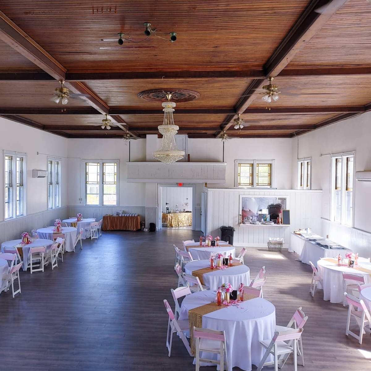 A spacious hall with round tables covered in white tablecloths and pink accents, arranged calmly around the room beneath a chandelier and wooden ceiling.