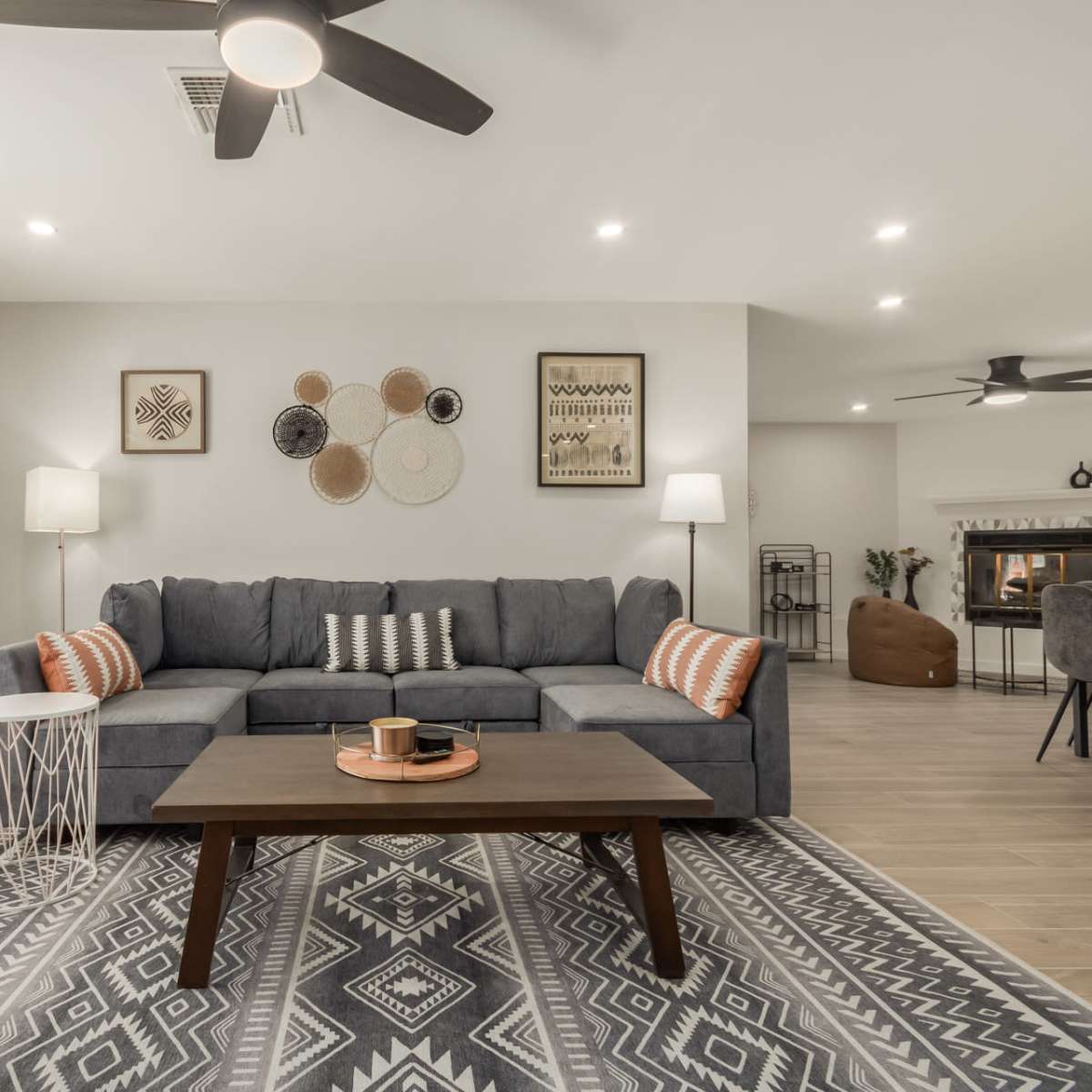A modern living room features a grey sectional sofa, a wooden coffee table, decorative wall hangings, and large windows allowing natural light.