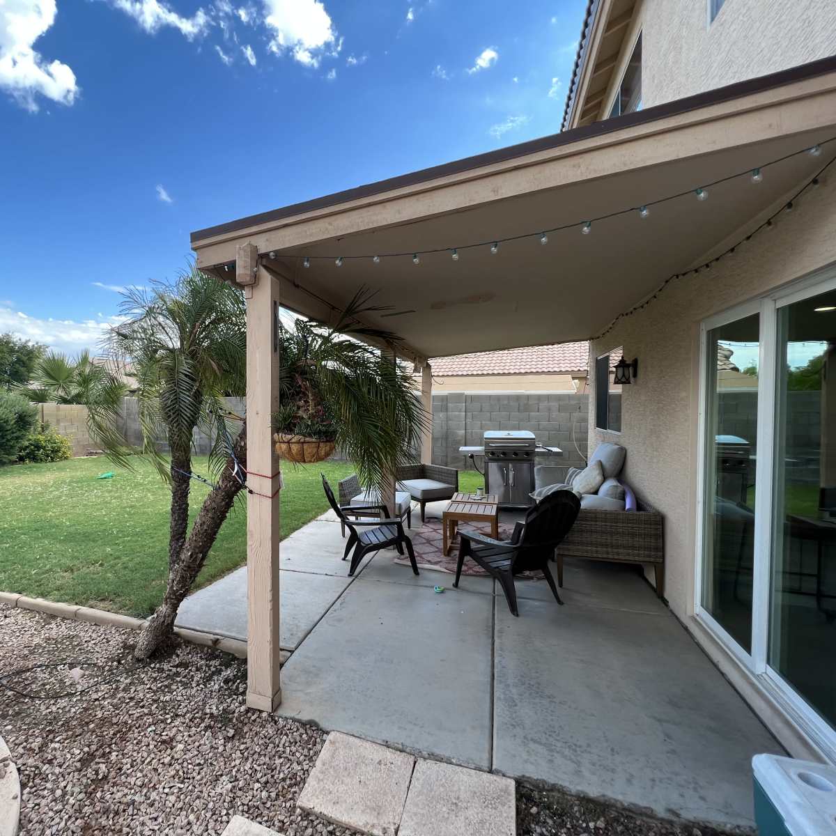 A patio area is shaded by a roof and features outdoor furniture, with a lawn and shrubs visible in the background.