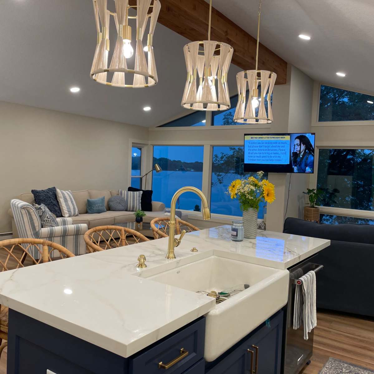 A modern kitchen with a marble countertop, a gold faucet, and a view of a living area featuring a sectional sofa and large windows overlooking a body of water.