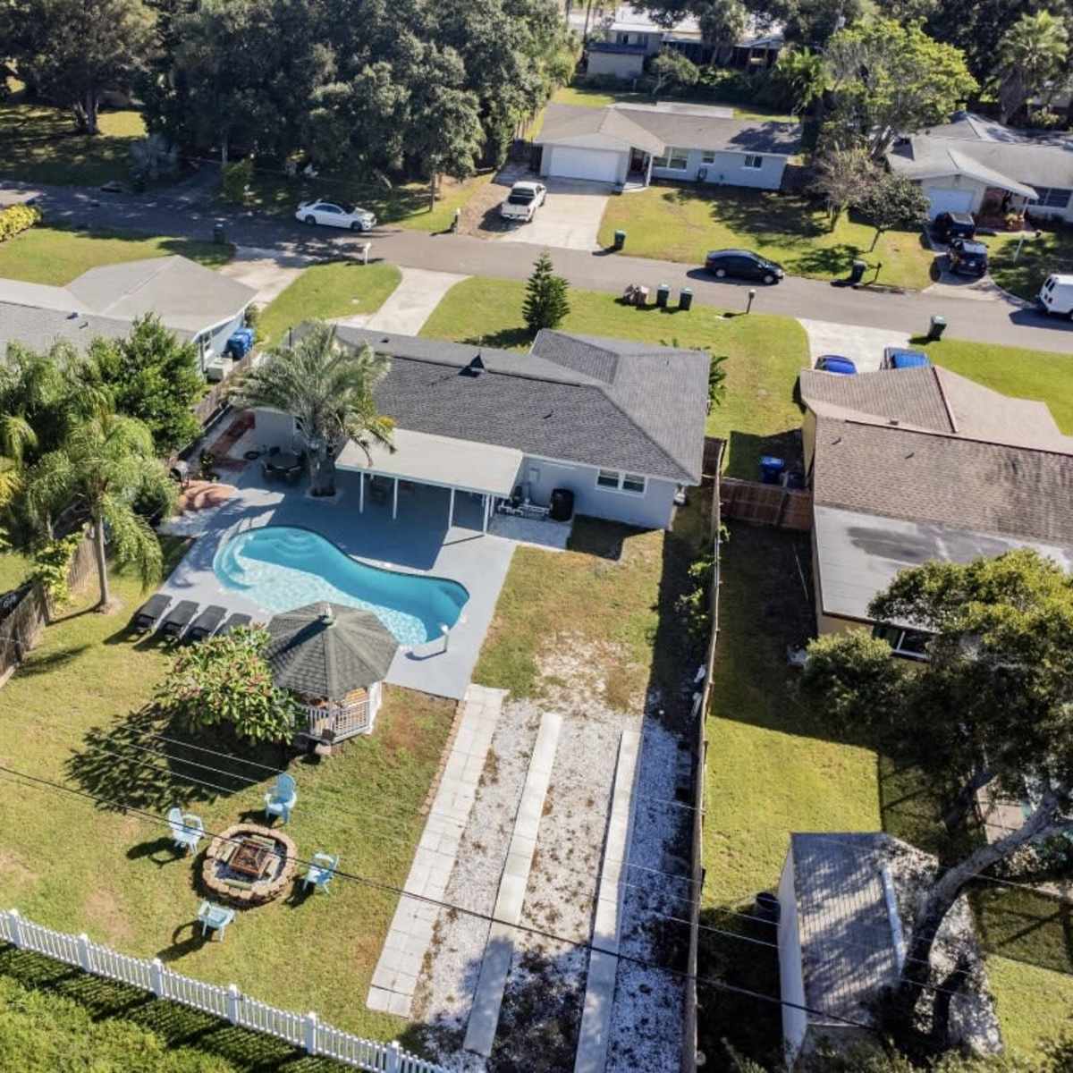 A residential backyard with a swimming pool, patio area, and a fire pit, surrounded by various trees and houses on a quiet street.