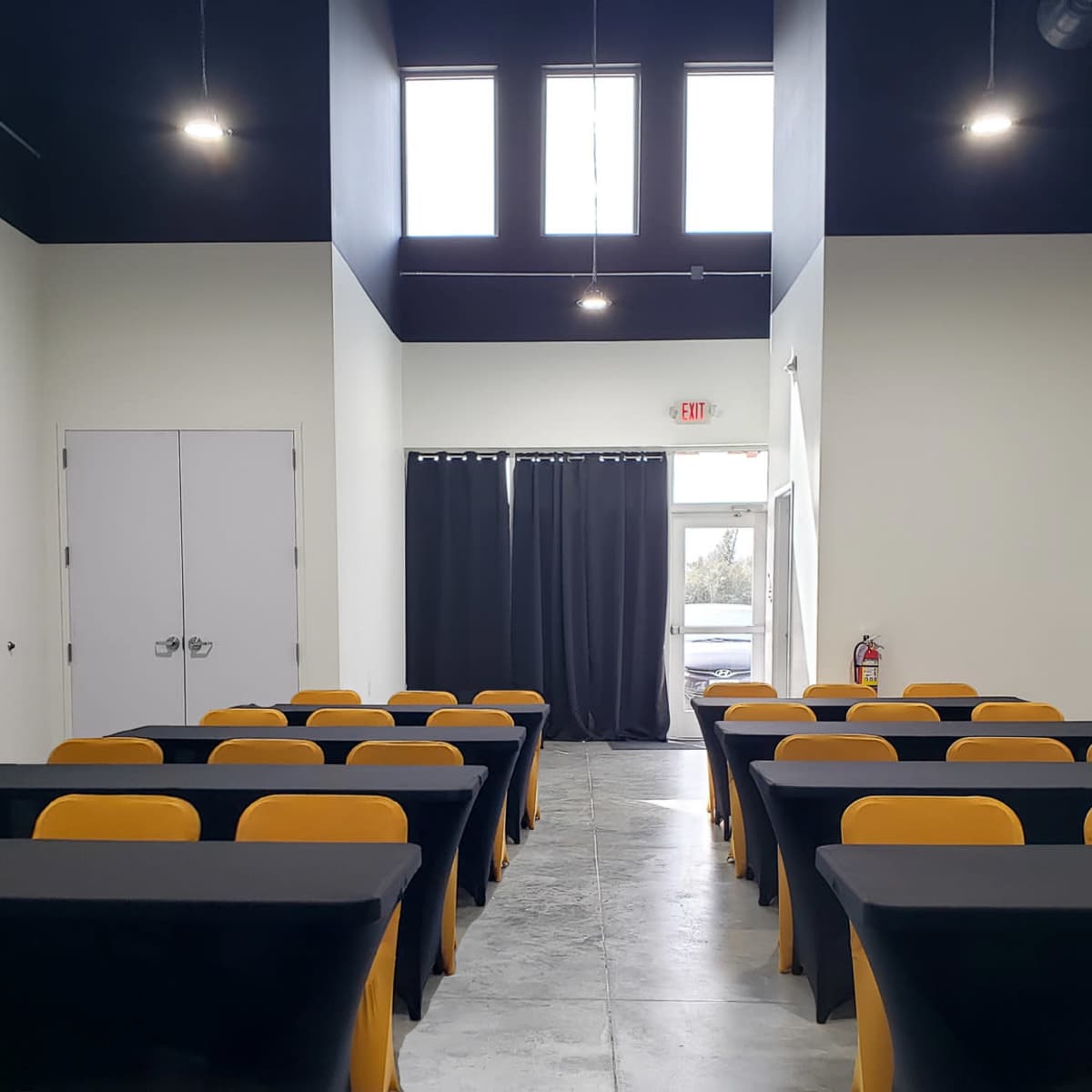 A modern conference room with rows of black tables and yellow chairs, lit by overhead lights and featuring a dark ceiling.