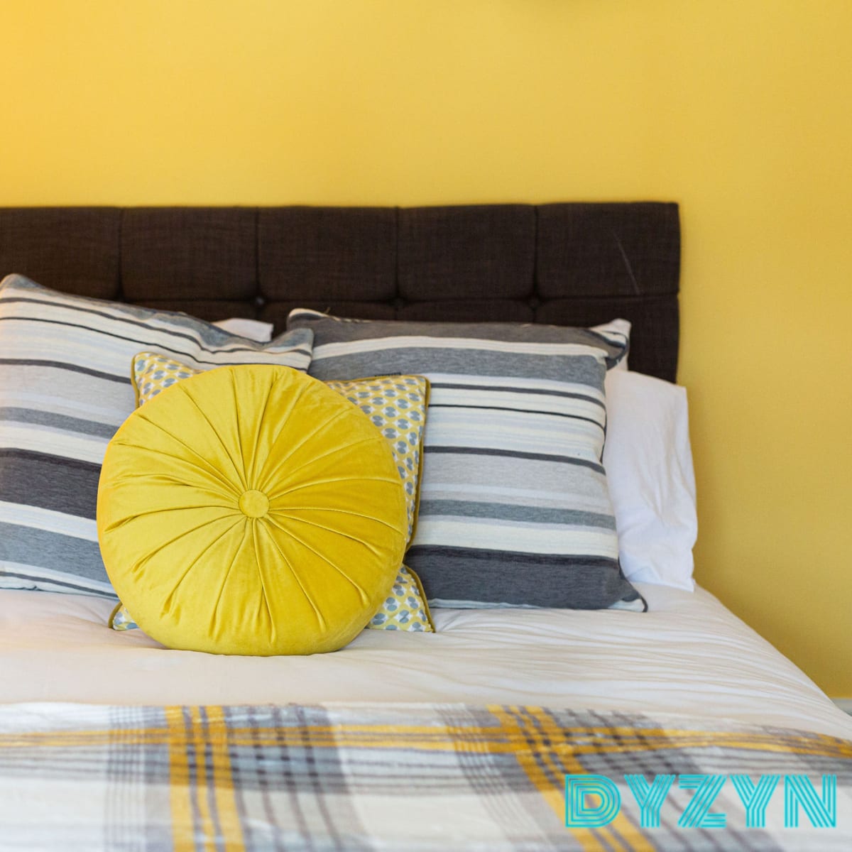 A neatly made bed with a dark headboard, striped pillows, and a large yellow round cushion against a bright yellow wall.