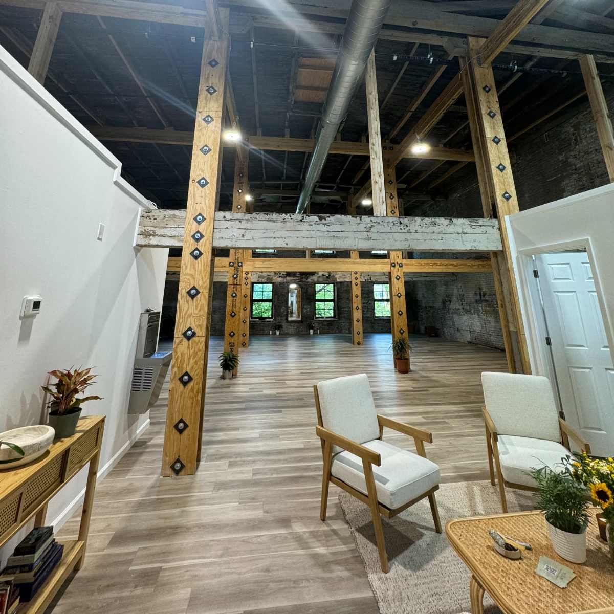 An interior space with wooden support beams, large windows, and minimal furniture, featuring a light-colored floor and exposed industrial elements.