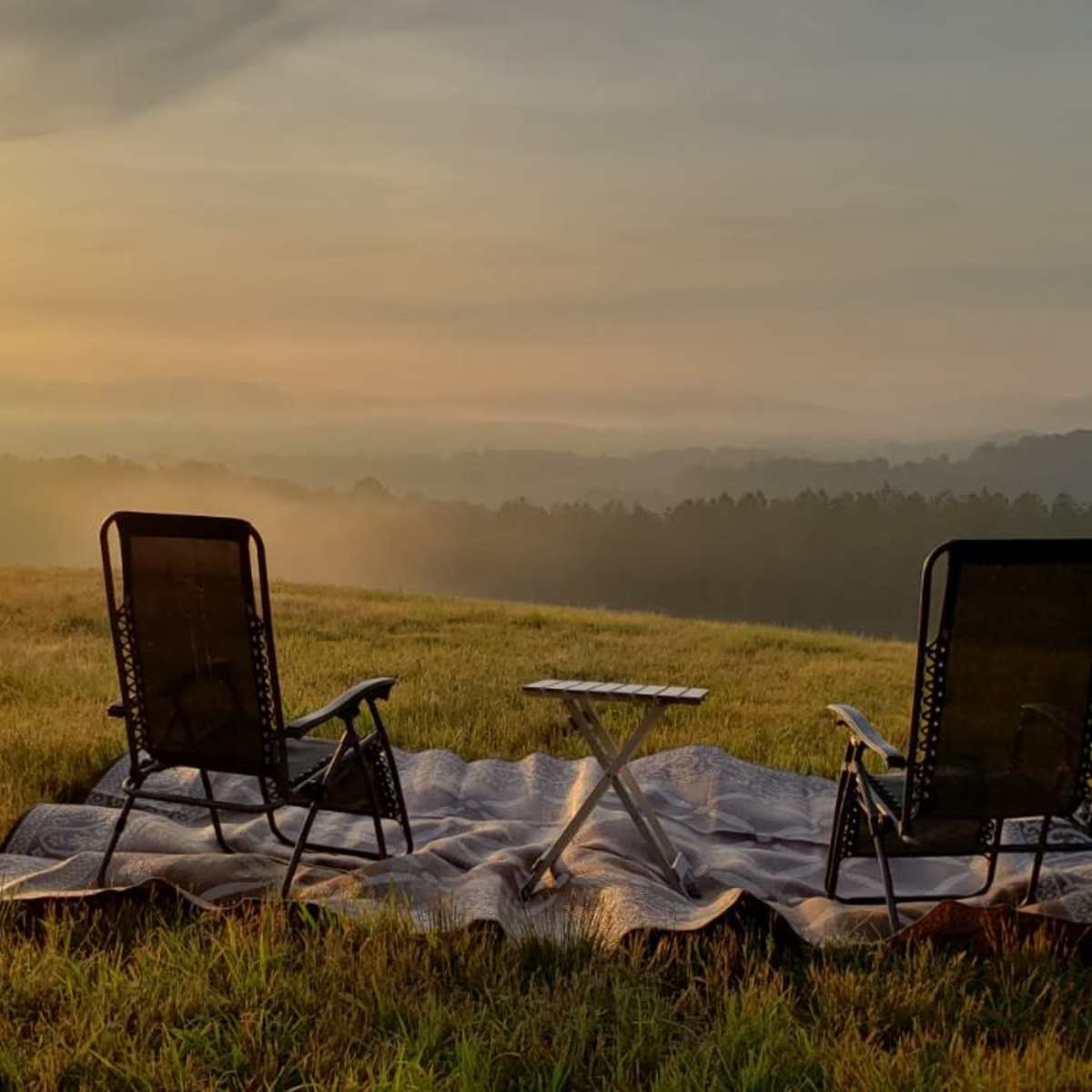 Two chairs are set up on a blanket in a grassy field, facing a sunrise over distant hills.