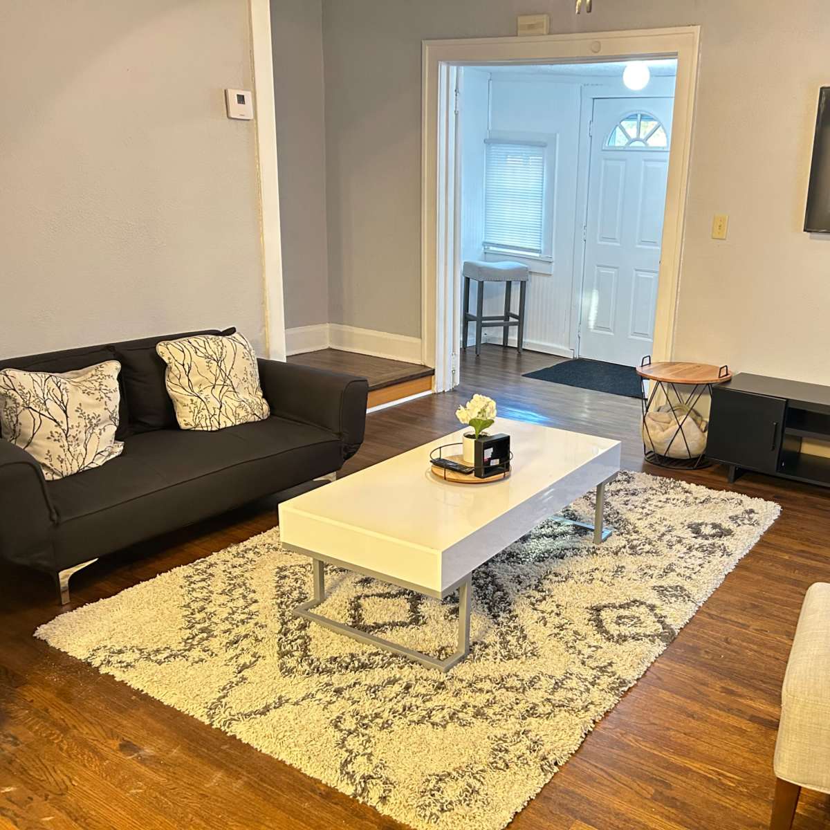 A living room with a black sofa, a white coffee table, a light gray armchair, and a TV mounted on the wall, all arranged on a patterned rug.