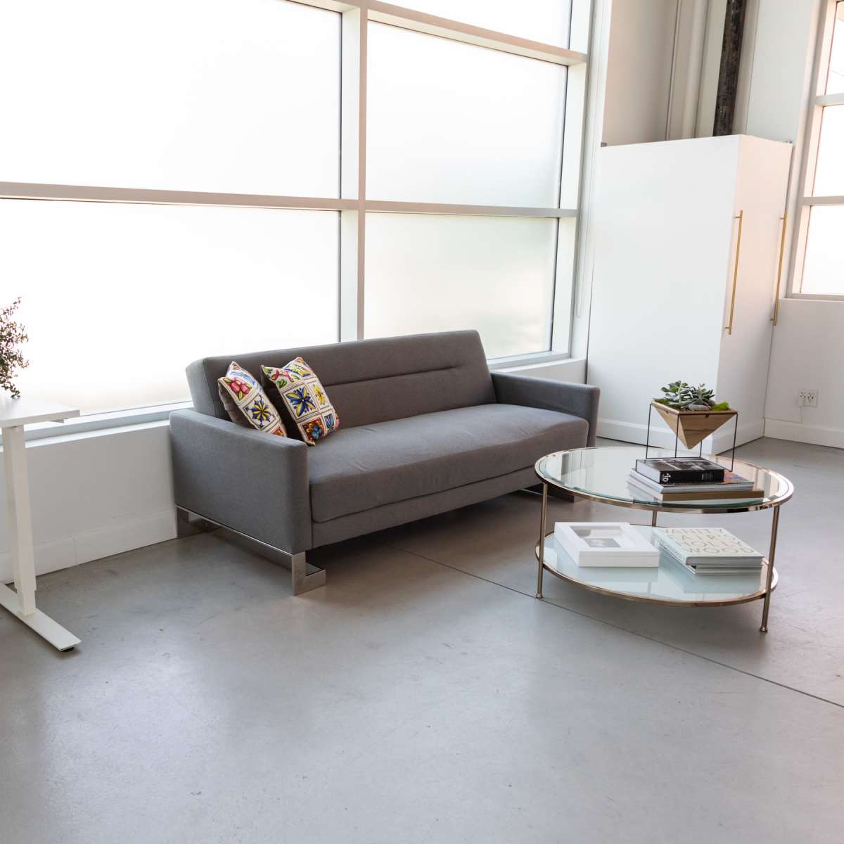 A modern, minimalist living space featuring a gray sofa, a glass coffee table, a white desk with a computer, and large windows allowing light to enter.