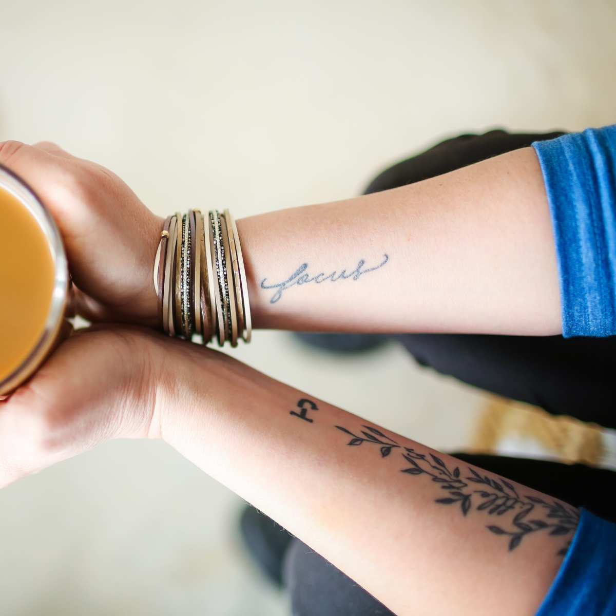 A person holds a glass mug filled with coffee, showcasing tattoos on their arms and several bracelets on one wrist.
