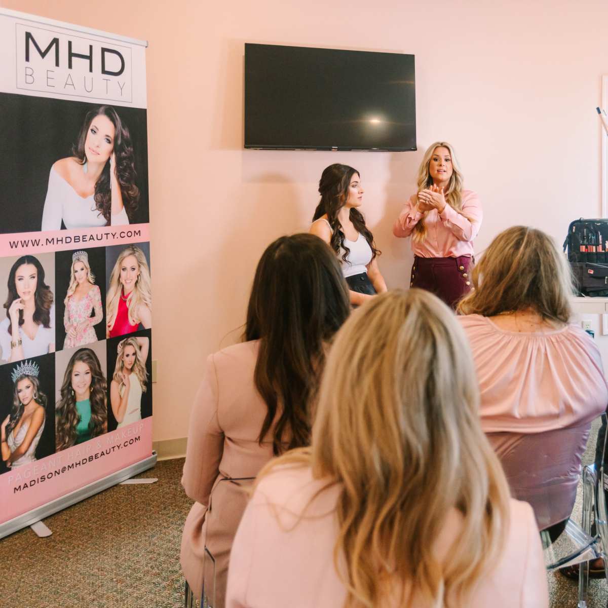 A speaker presents to an audience of women at a beauty workshop, with a promotional banner for MHD Beauty in the background.