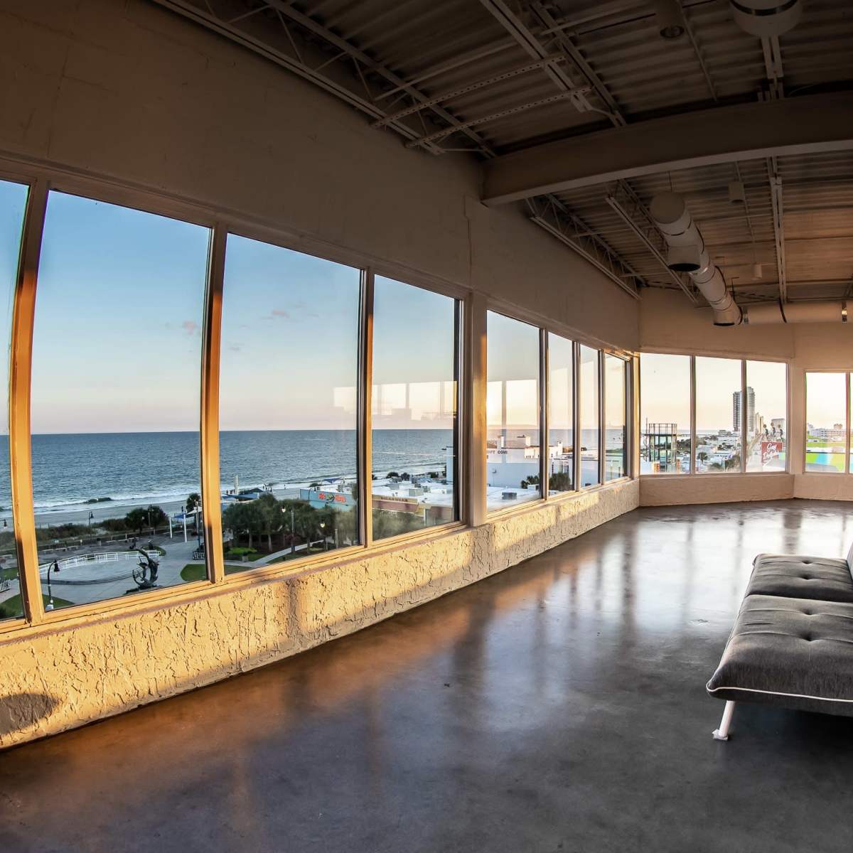 A spacious room with large windows offers a view of the ocean and shoreline at sunset.