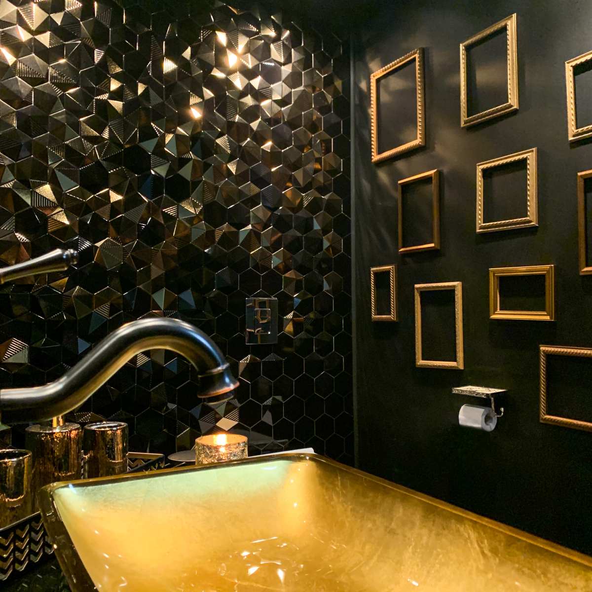 A modern bathroom features a hexagonal black tile wall, a gold-colored sink, and several empty, framed outlines on a contrasting wall.