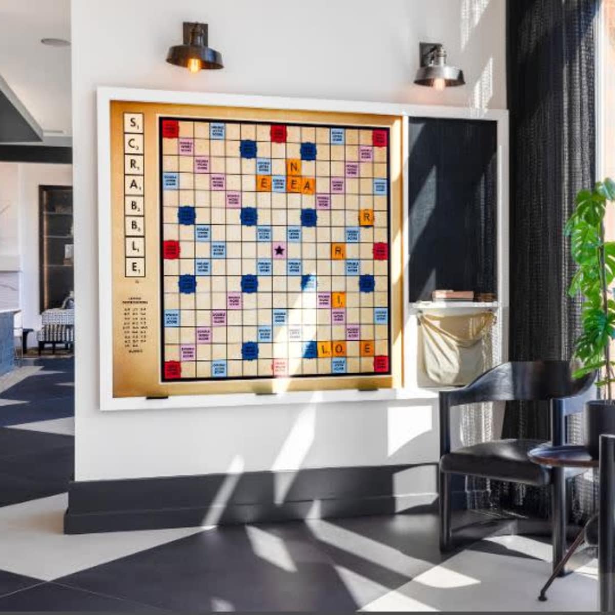 A Scrabble board is displayed on a wall in a modern seating area with large windows, a potted plant, and a black chair.