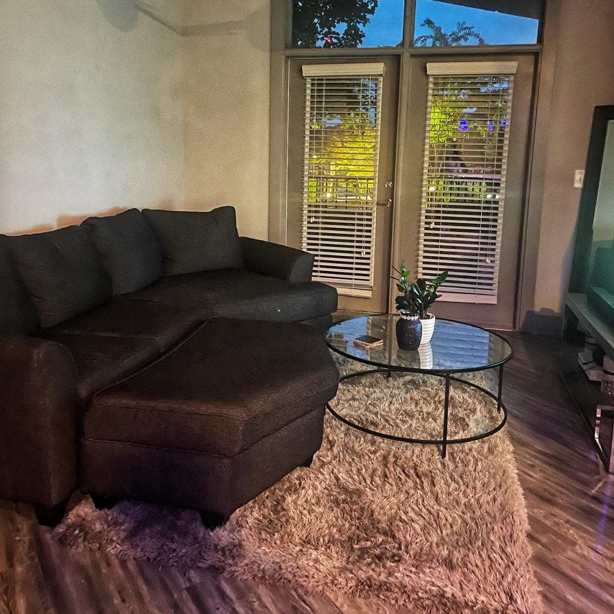A dark sectional sofa and a glass coffee table sit on a rug in a well-lit living room with large sliding doors.