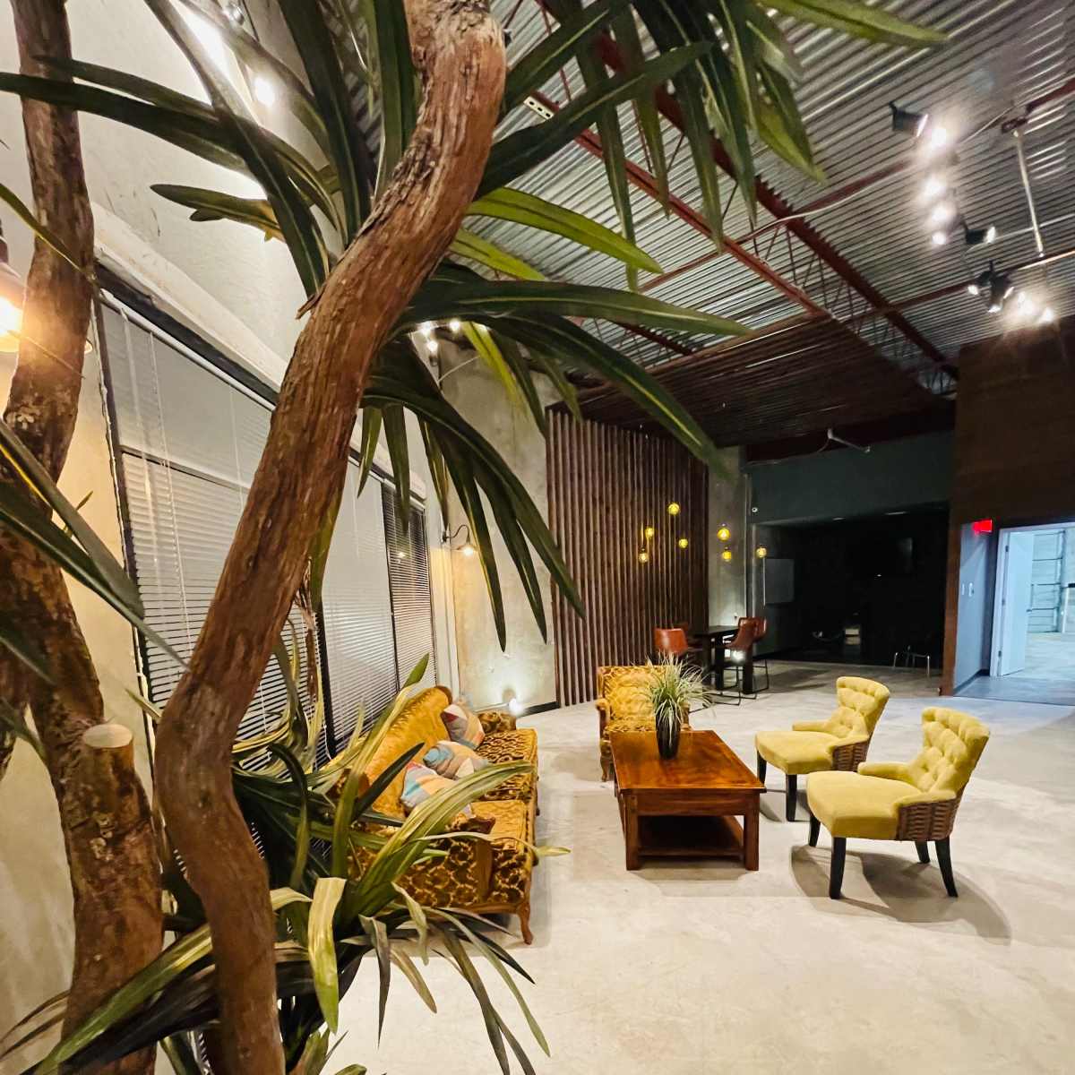 A modern interior with a sofa and yellow chairs arranged around a wooden coffee table under soft lighting, surrounded by potted plants.