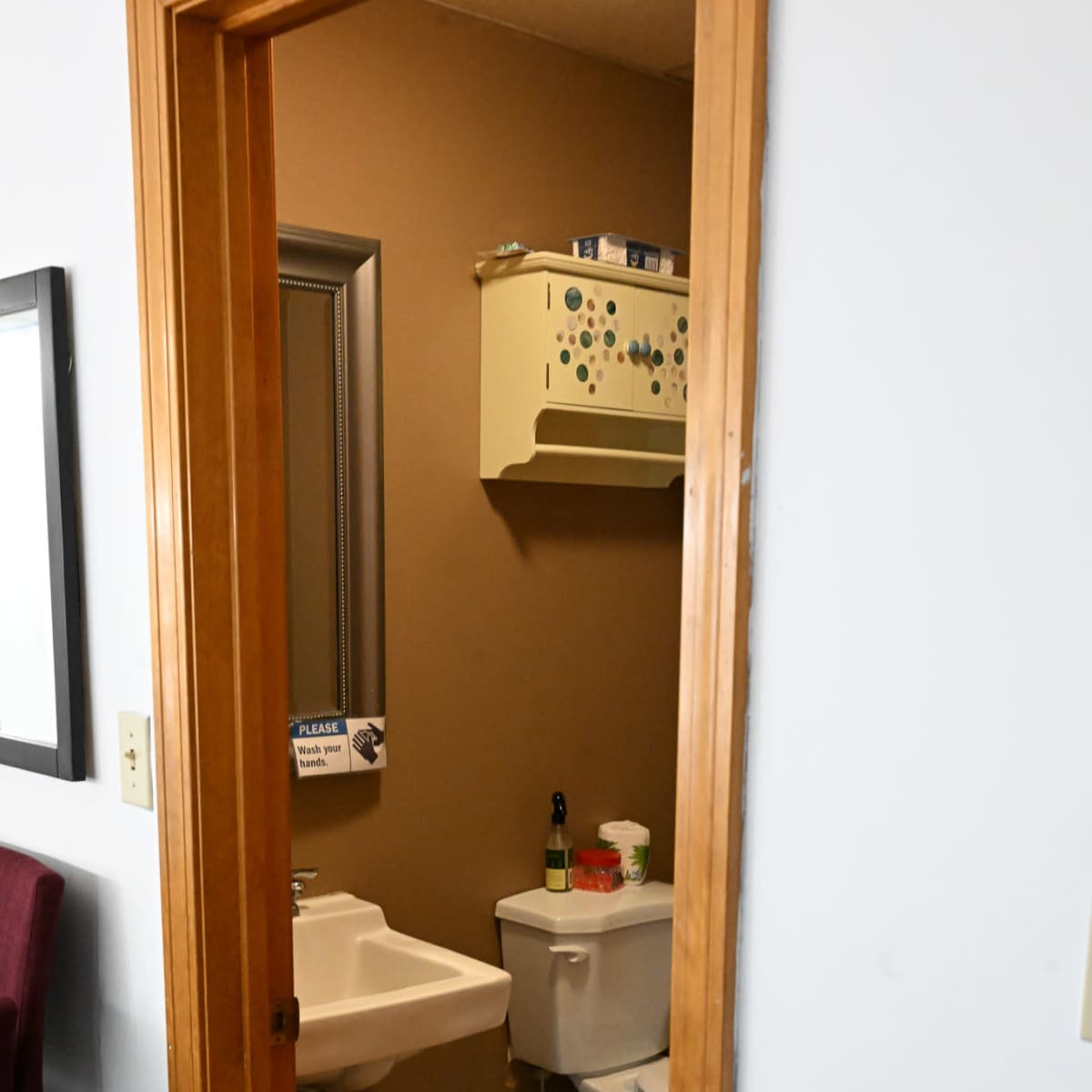 A small bathroom accessed through a wooden door, featuring a sink, a toilet, and a shelf with various items.
