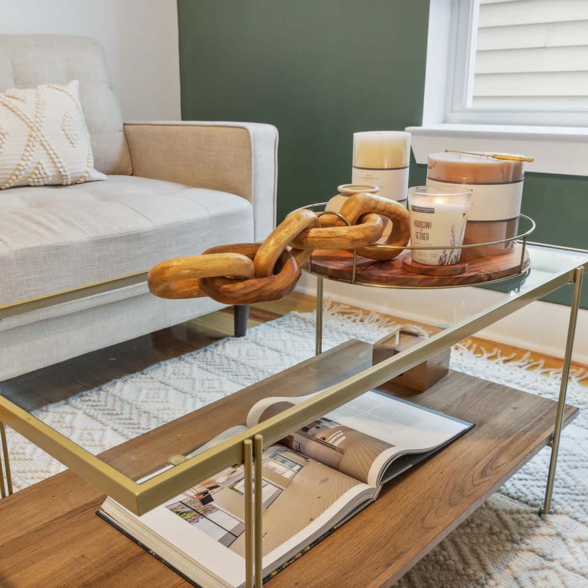 A modern living room scene features a beige sofa with decorative cushions, a glass and gold coffee table holding a wooden chain sculpture and candles, and a magazine on a wooden shelf below.