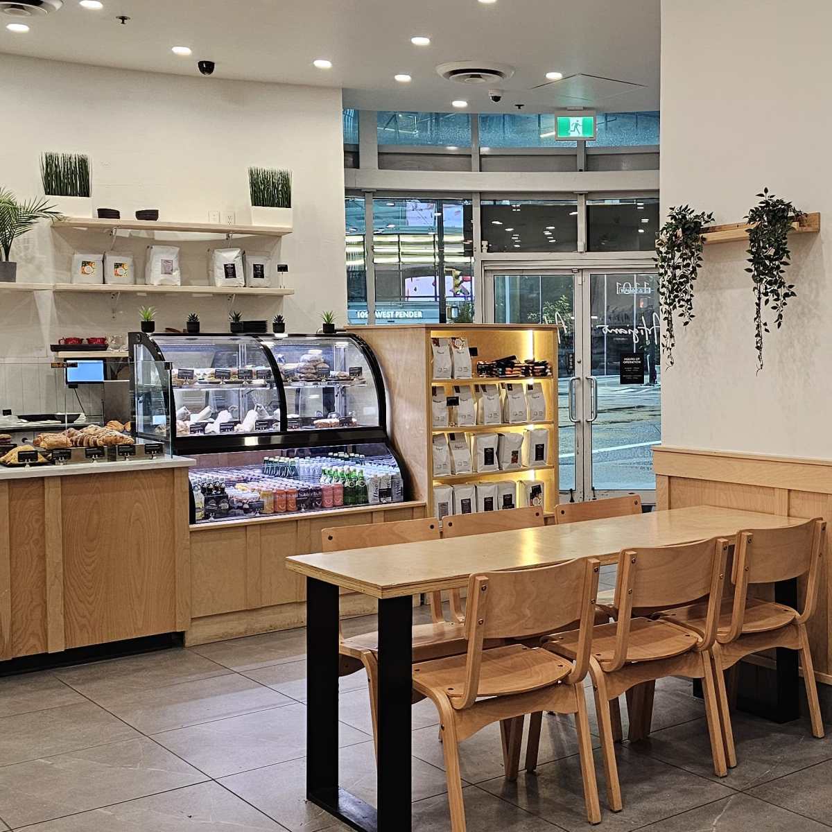 A bright café features wooden tables and chairs, a display case with food items, and large windows showing an outdoor seating area.