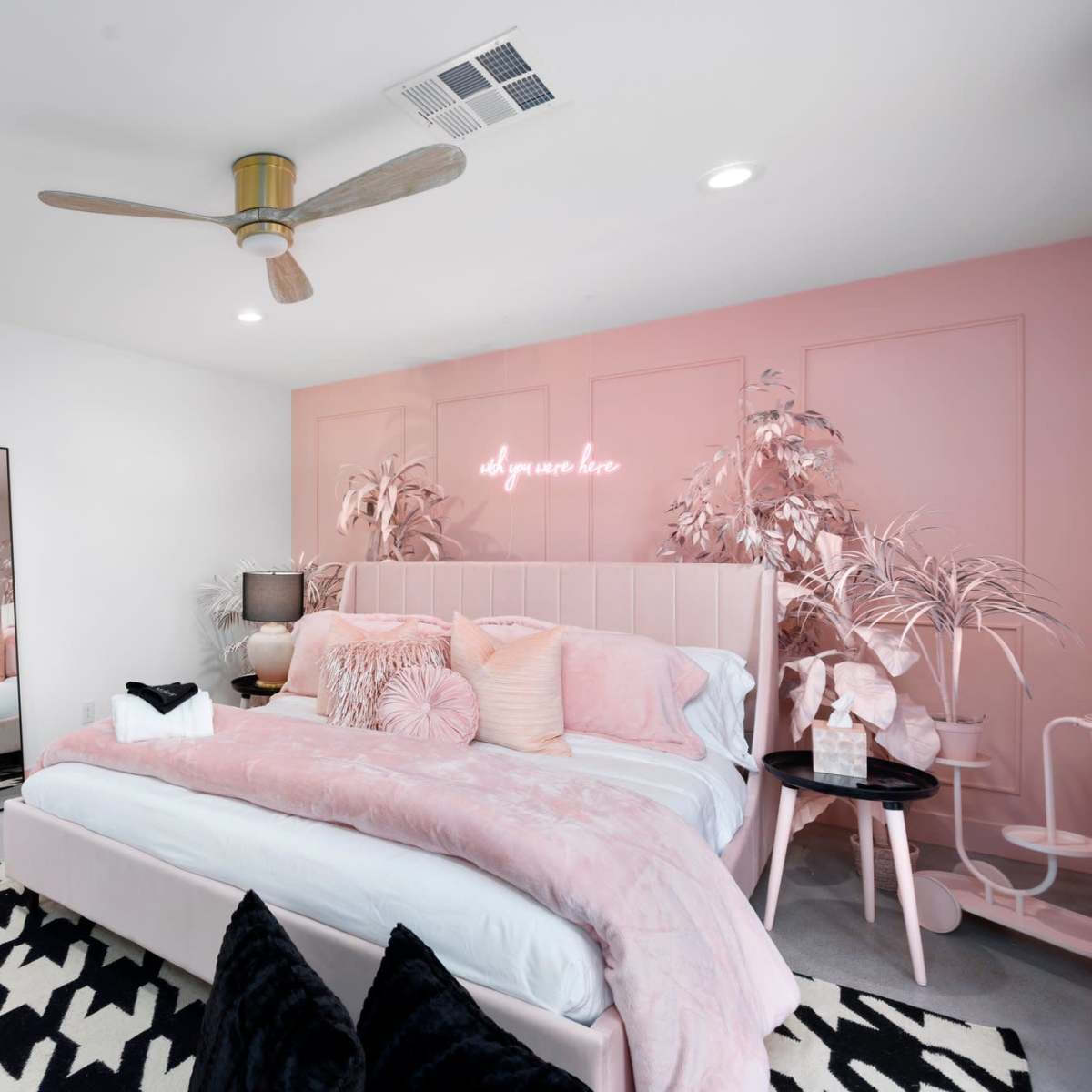 A modern bedroom with a pink and white color scheme, featuring a large bed, a mirror, a ceiling fan, and a decorative rug.