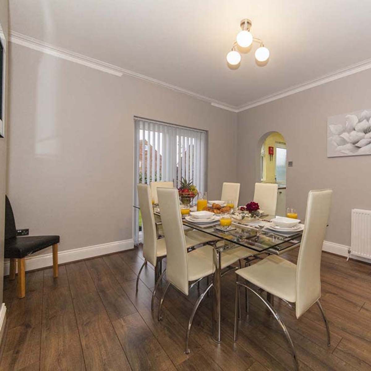 A dining room features a glass table set for a meal with six chairs, a heater on the wall, and a large wall decoration of a flower.