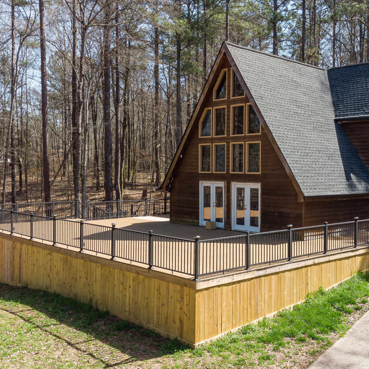 A brown A-frame house with large windows is surrounded by trees, featuring a large deck and a paved driveway.