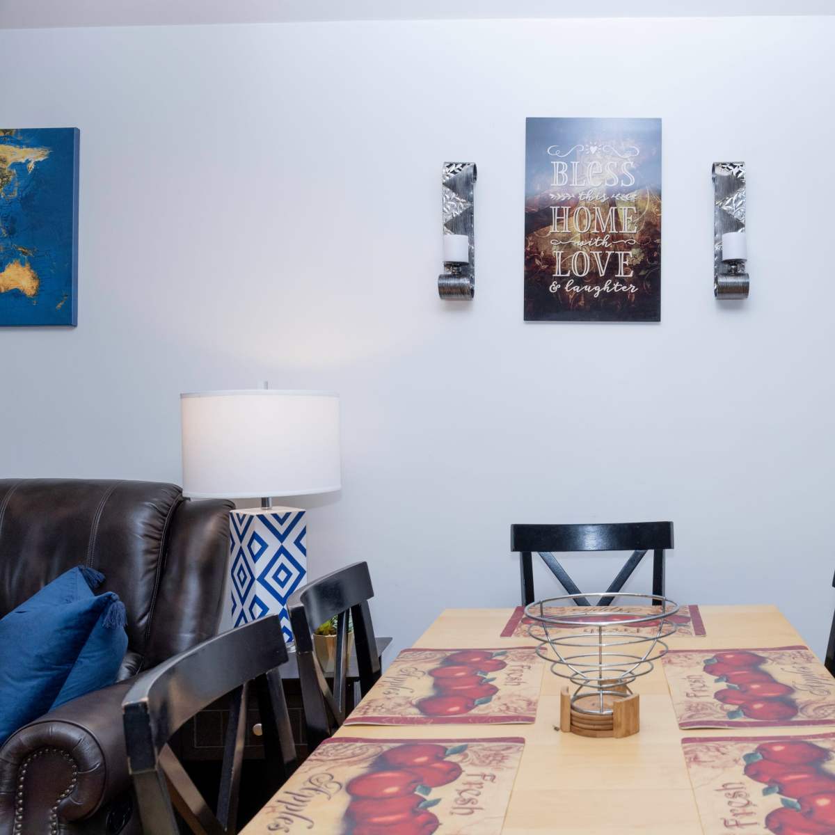 A dining area with a wooden table set with a fruit-themed tablecloth, surrounded by black chairs, and wall decorations above.
