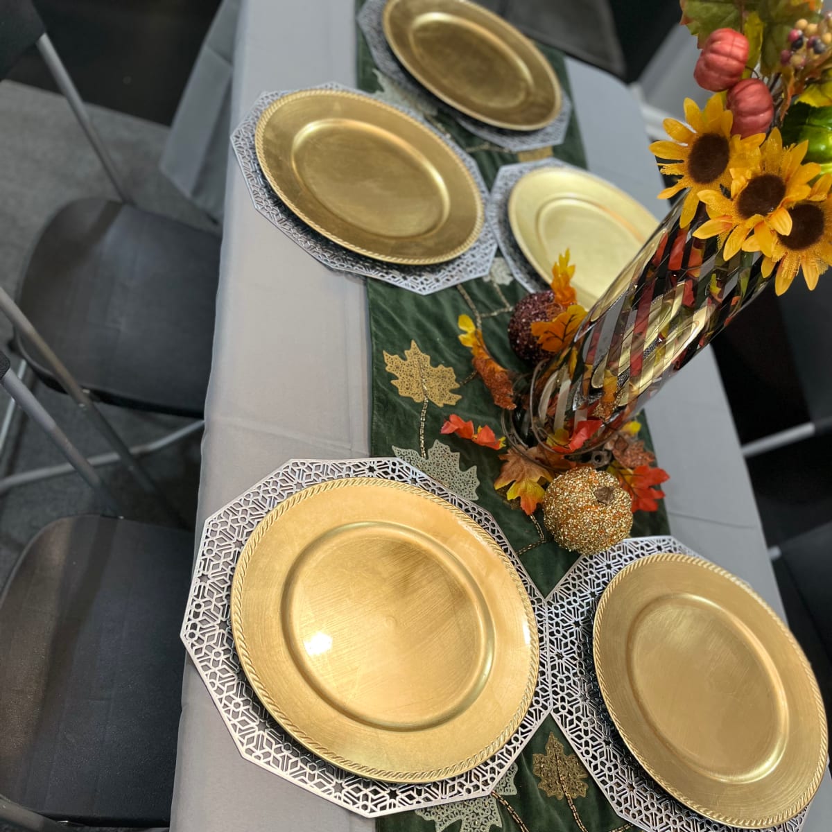 A dining table set with gold plates, a green table runner adorned with leaves, and a floral centerpiece featuring sunflowers and decorative items.
