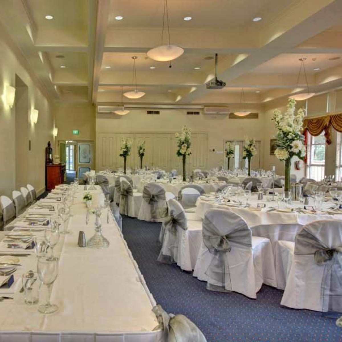 A spacious banquet hall is set with elegantly arranged tables covered in white cloths and adorned with floral centerpieces.