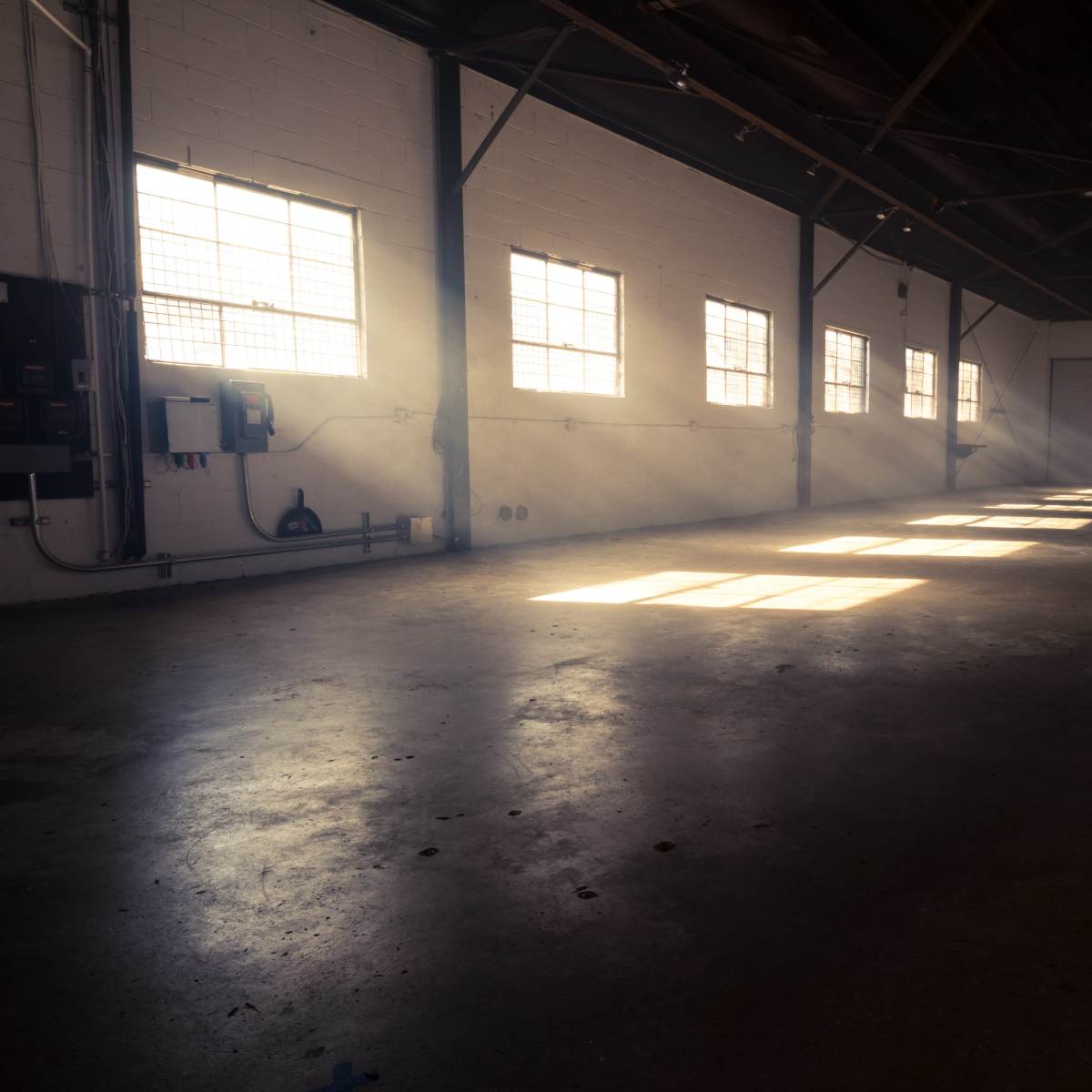 A sunlit industrial space features an empty concrete floor and large windows casting shadows along the walls.