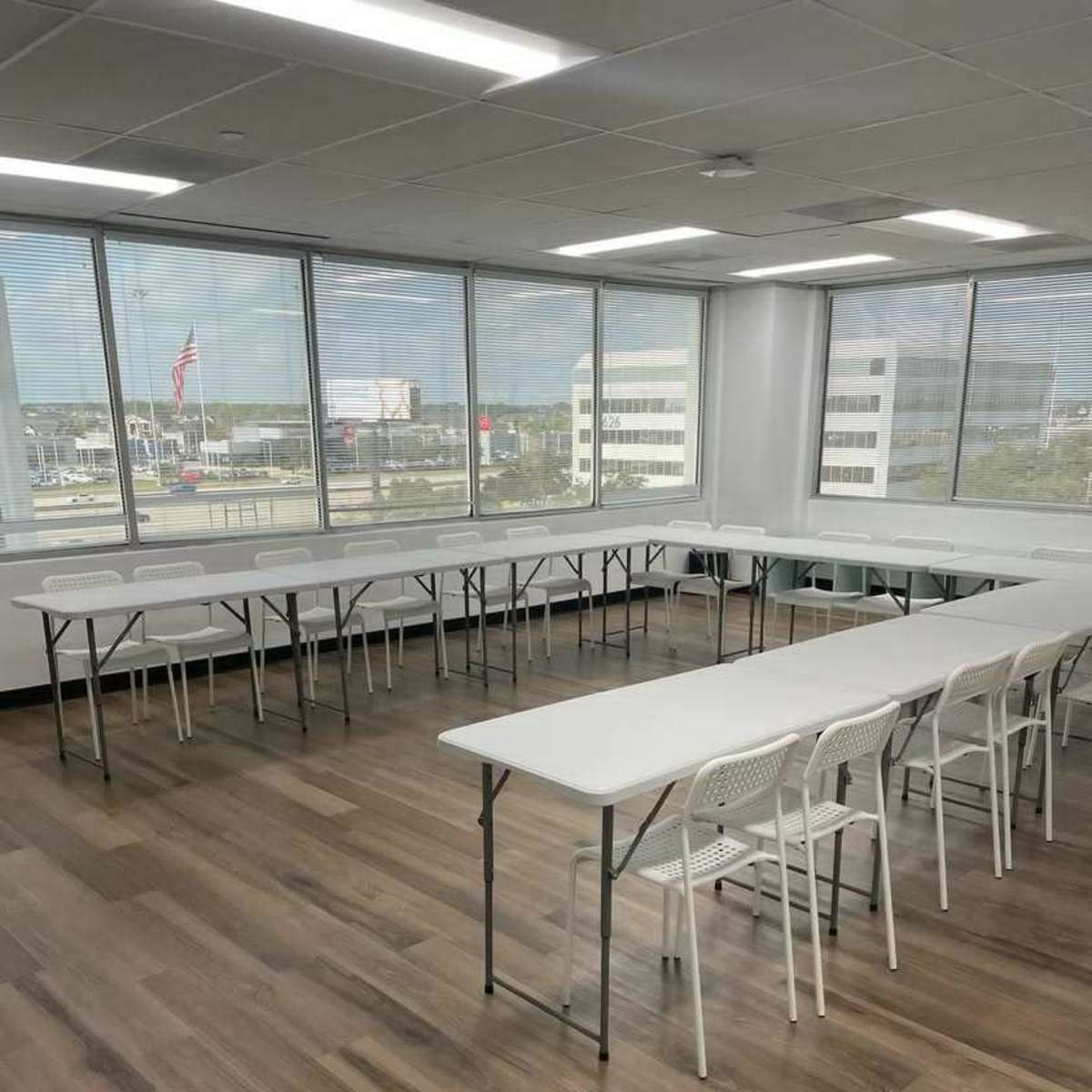 A well-lit meeting room features several long tables arranged in a U-shape, surrounded by white chairs, with large windows showing an urban view.