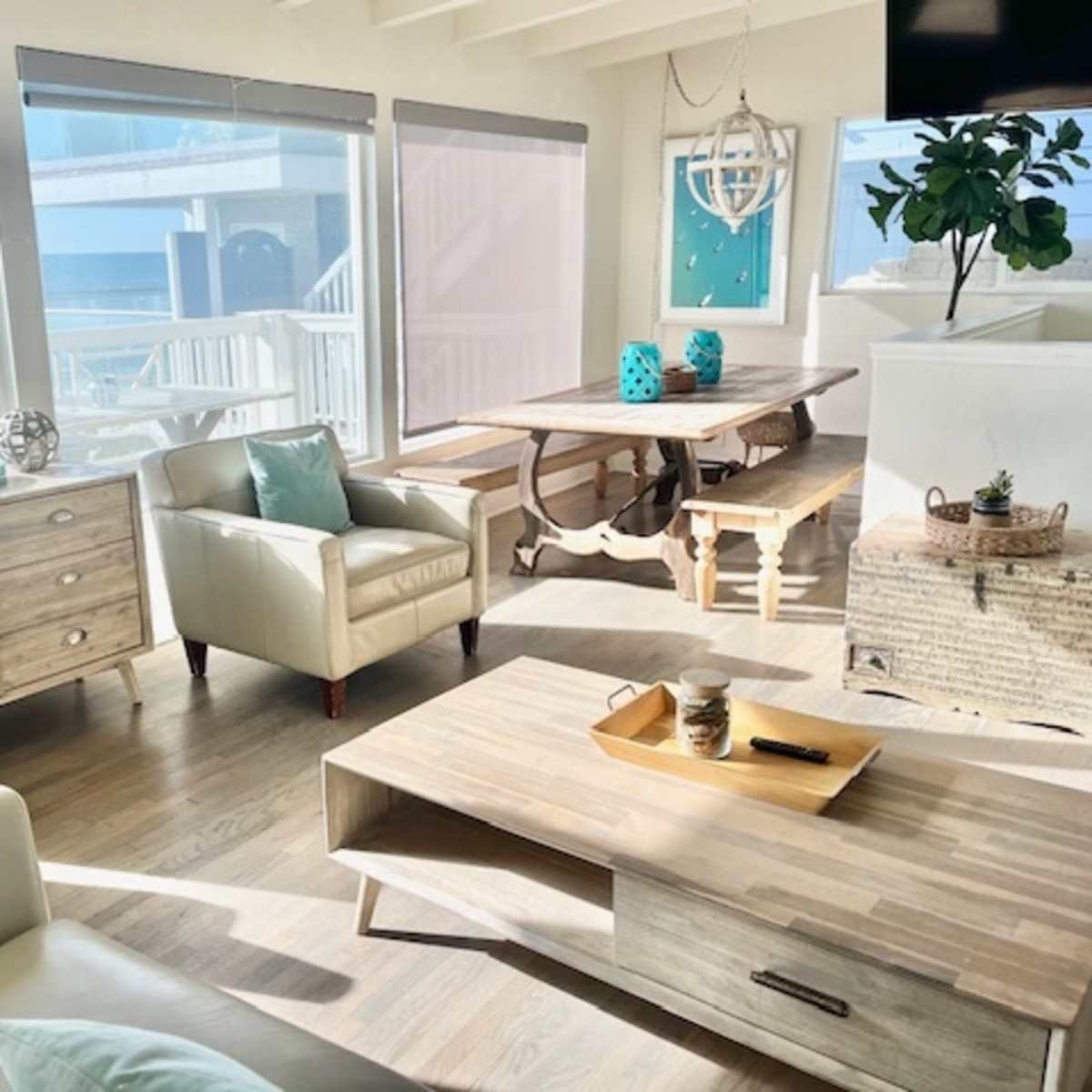 A bright coastal living room with large windows overlooking the ocean, featuring light-colored furniture and a wooden dining table.